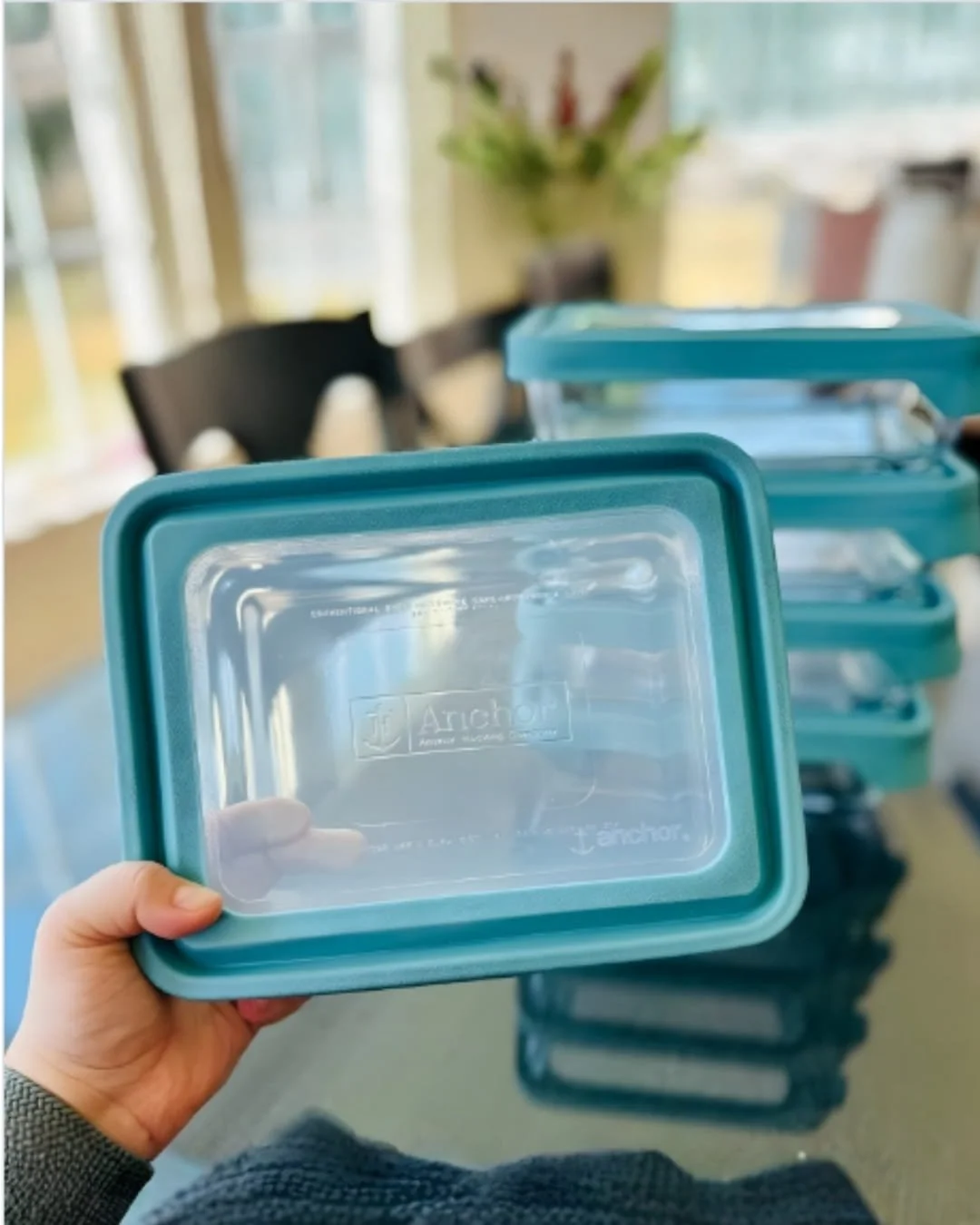 Teal-rimmed Anchor Hocking food storage lid with a clear center, held by a hand, with a stack of matching glass containers in the background, set in a brightly lit kitchen or dining area.