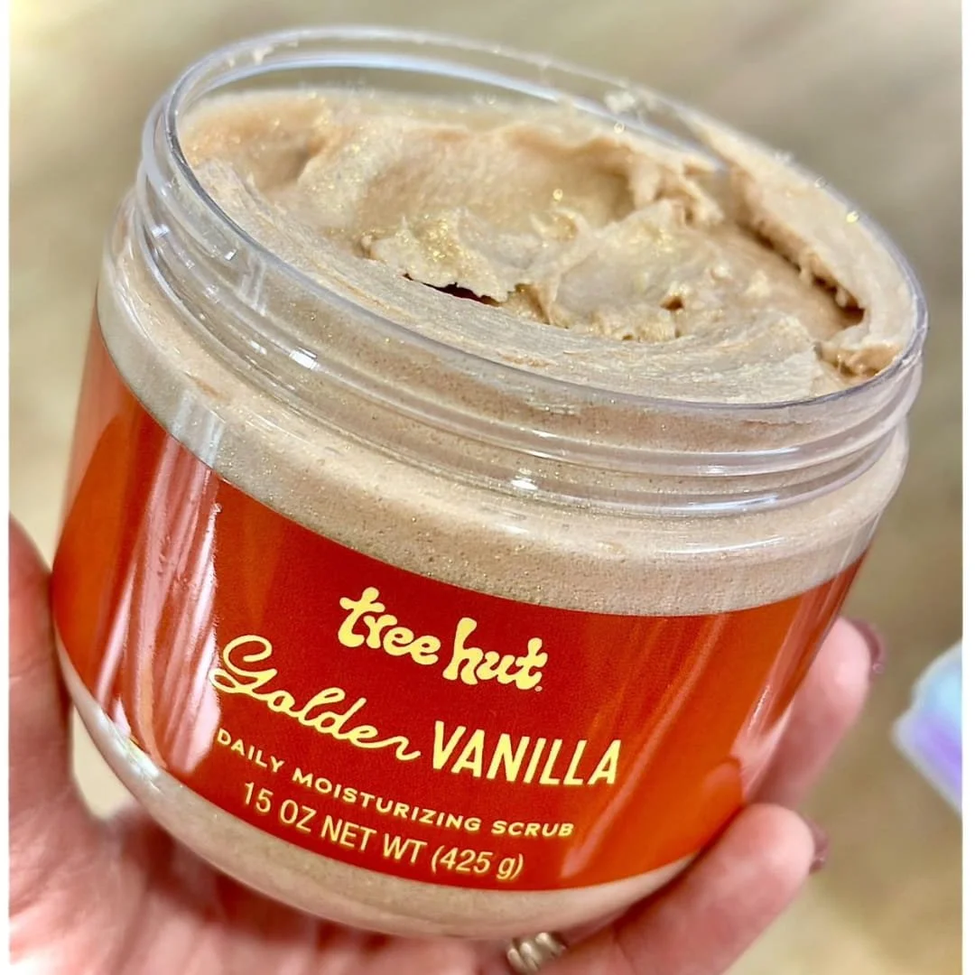 Tree Hut Golden Vanilla Daily Moisturizing Scrub in a clear jar with orange label, showcasing a light brown creamy texture with golden shimmer, held by a hand against a warm-toned background.