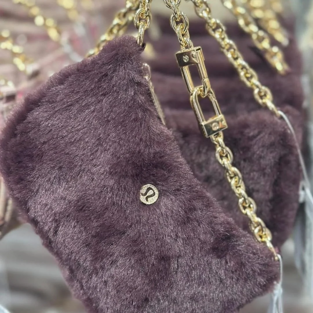 Lululemon faux-fur shoulder bag in deep purple with a gold chain strap and circular gold logo, displayed against a blurred retail background.
