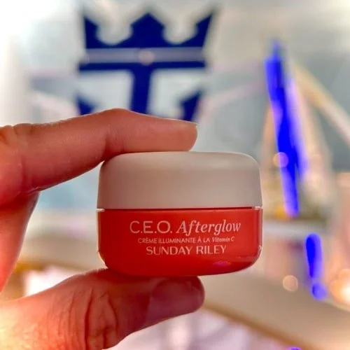 Hand holding an orange jar of Sunday Riley C.E.O. Afterglow Vitamin C cream with a white lid, set against a blurred background featuring a blue Royal Caribbean logo and abstract glowing elements.