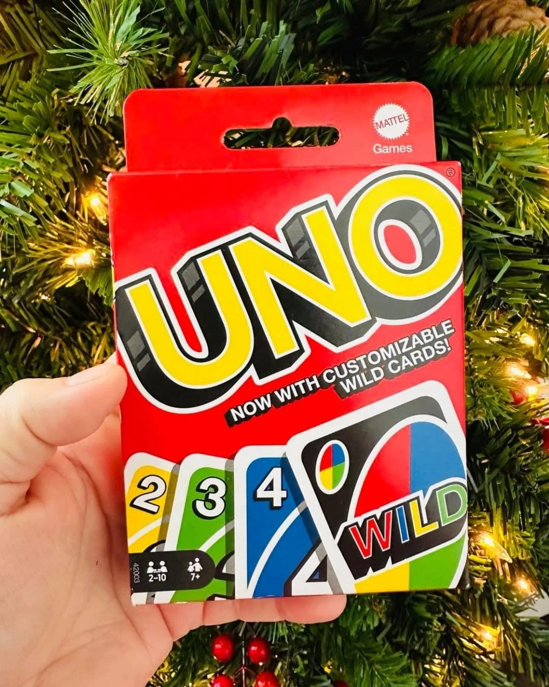 Box of Mattel's UNO card game featuring customizable wild cards, held in a hand against a festive Christmas tree background with warm lights and red ornaments.