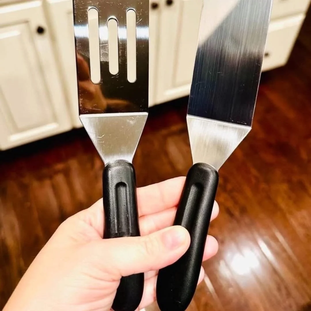 Two small stainless steel spatulas with black plastic handles, one slotted and one solid, against a kitchen background with light cabinets and a dark wooden floor