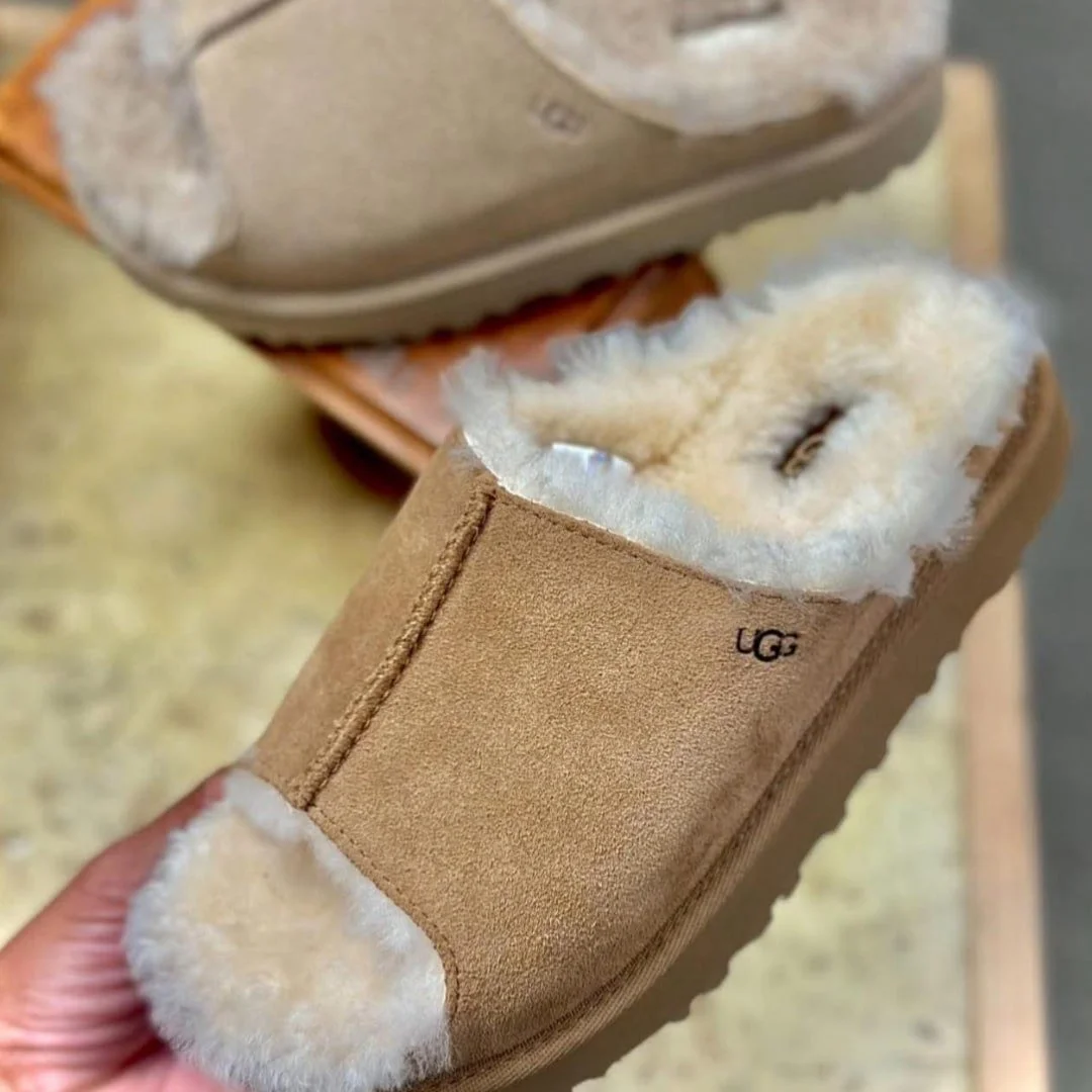 UGG tan suede slip-on slippers with cream sheepskin lining, featuring a ribbed sole and visible UGG logo, displayed on a light wooden surface.