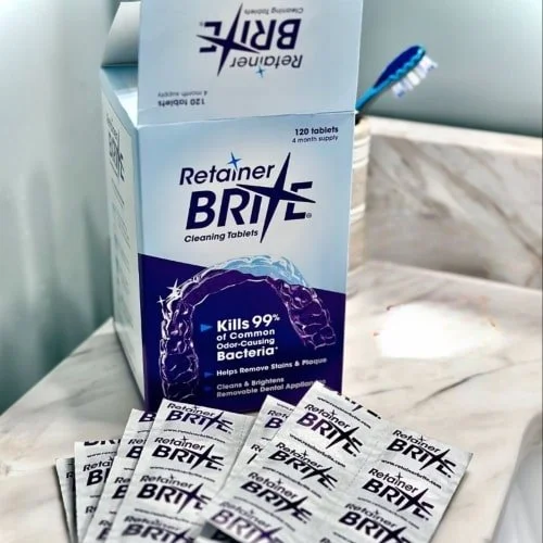 Box of Retainer BRITE Cleaning Tablets with 120 tablets for a 4-month supply, shown slightly open with individual silver foil packets fanned out in front, set on a white surface with a blue toothbrush in the background.