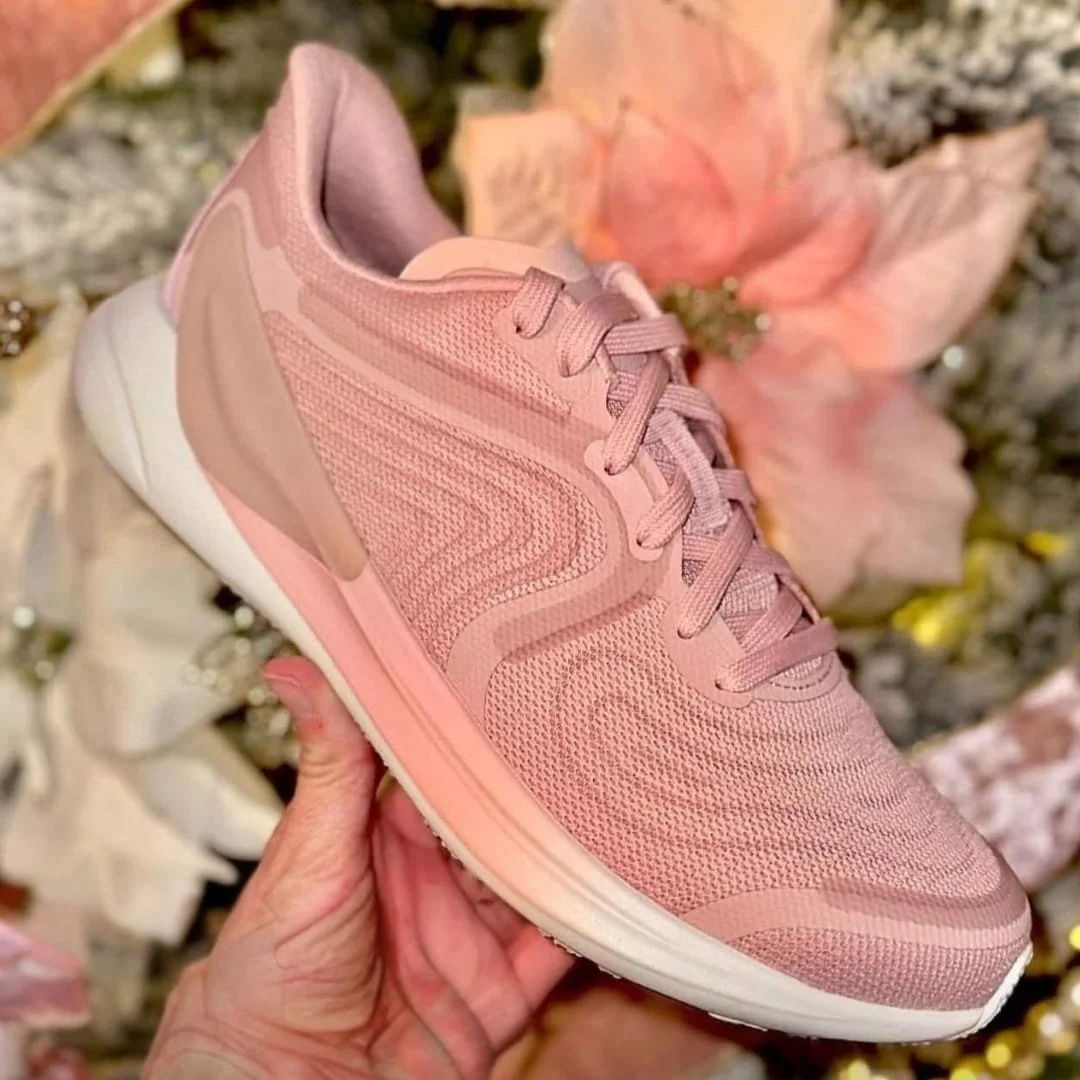 Light pink Women's Blissfeel running shoe with a mesh upper, matching laces, and a two-tone sole, held by a hand against a blurred festive background with soft pink and white decor.