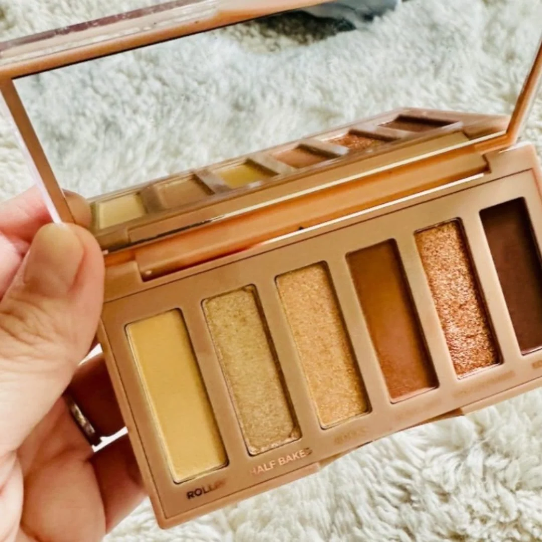 Open mini eyeshadow palette with six neutral matte shades and a built-in mirror, displayed on a textured surface beside a small potted plant