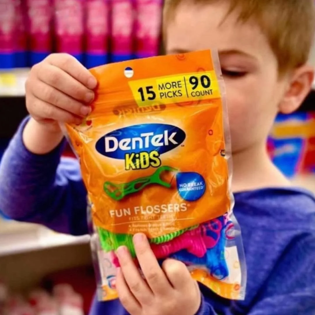 Child holding a package of DenTek Kids Fun Flossers with colorful flossers visible inside, featuring 90-count, no-break guarantee, and fun design for kids, in a retail store setting.