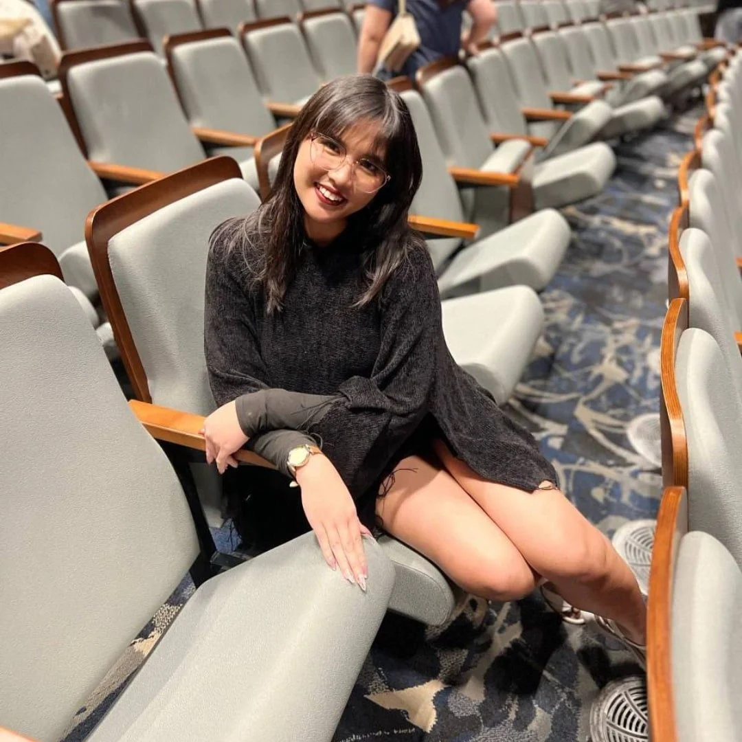 Khim, content creator for Nicole Fabulous Finds, seated in an empty auditorium wearing glasses, a dark sweater, and white sneakers, with rows of gray seats and patterned carpet in the background.