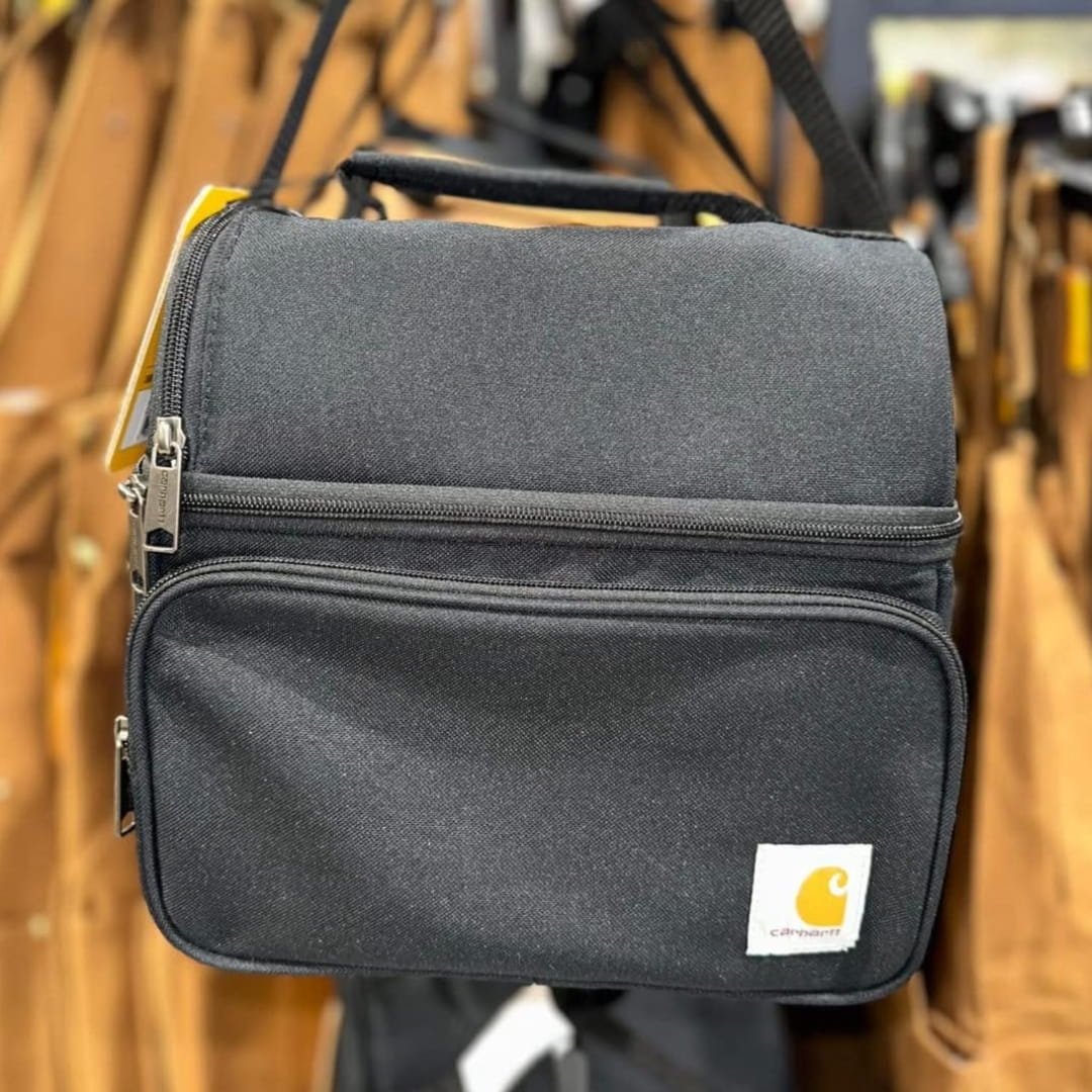 Carhartt black lunch cooler bag with dual zippered compartments, top handle, and shoulder strap, featuring the iconic Carhartt logo, displayed in a retail setting.