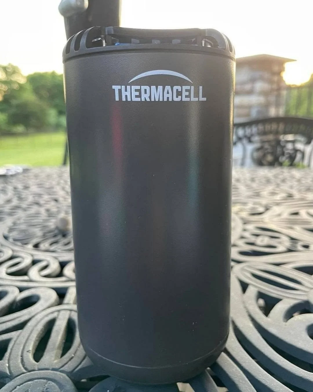 Black Thermacell mosquito repeller standing upright on a decorative outdoor metal table with a blurred lawn and patio setting in the background