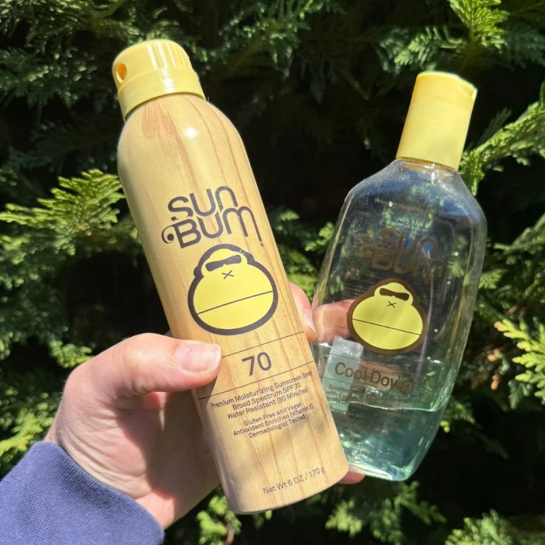 Two Sun Bum products—a Premium Moisturizing Sunscreen Spray SPF 70 with a wood-grain design and a Cool Down aloe gel in a clear bottle—held in a hand against a green leafy outdoor background.