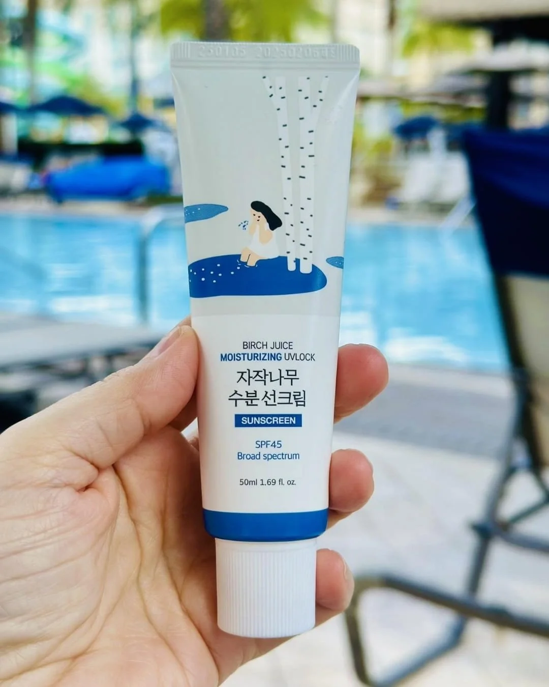 Hand holding a 50ml tube of Birch Juice Moisturizing Sunscreen with SPF45, featuring a whimsical illustration of a woman by birch trees and water, set against a blurred background of a swimming pool and tropical greenery.