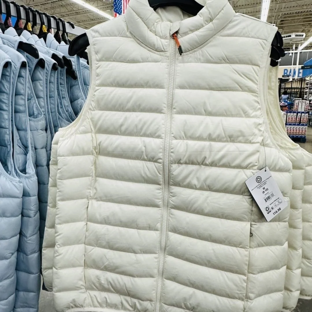 White sleeveless women's puffer vest with horizontal quilting and orange zipper pull, displayed on a hanger in a retail store with similar vests in the background.