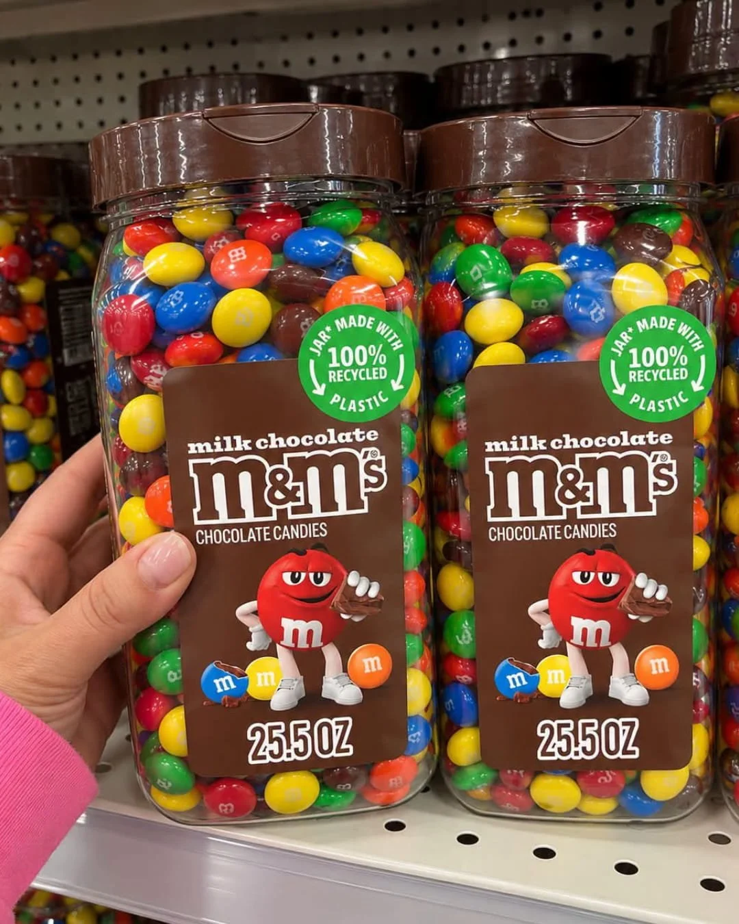 Hand holding a jar of M&M'S milk chocolate candy on a store shelf next to another 25.5 ounce jar