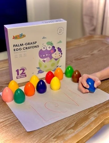 Product display of JupTup Palm-Grasp Egg Crayons for toddlers, featuring a box with a playful dinosaur illustration, 11 colorful egg-shaped crayons on white paper, and a child's hand drawing LOVE with a blue crayon on a wooden table.