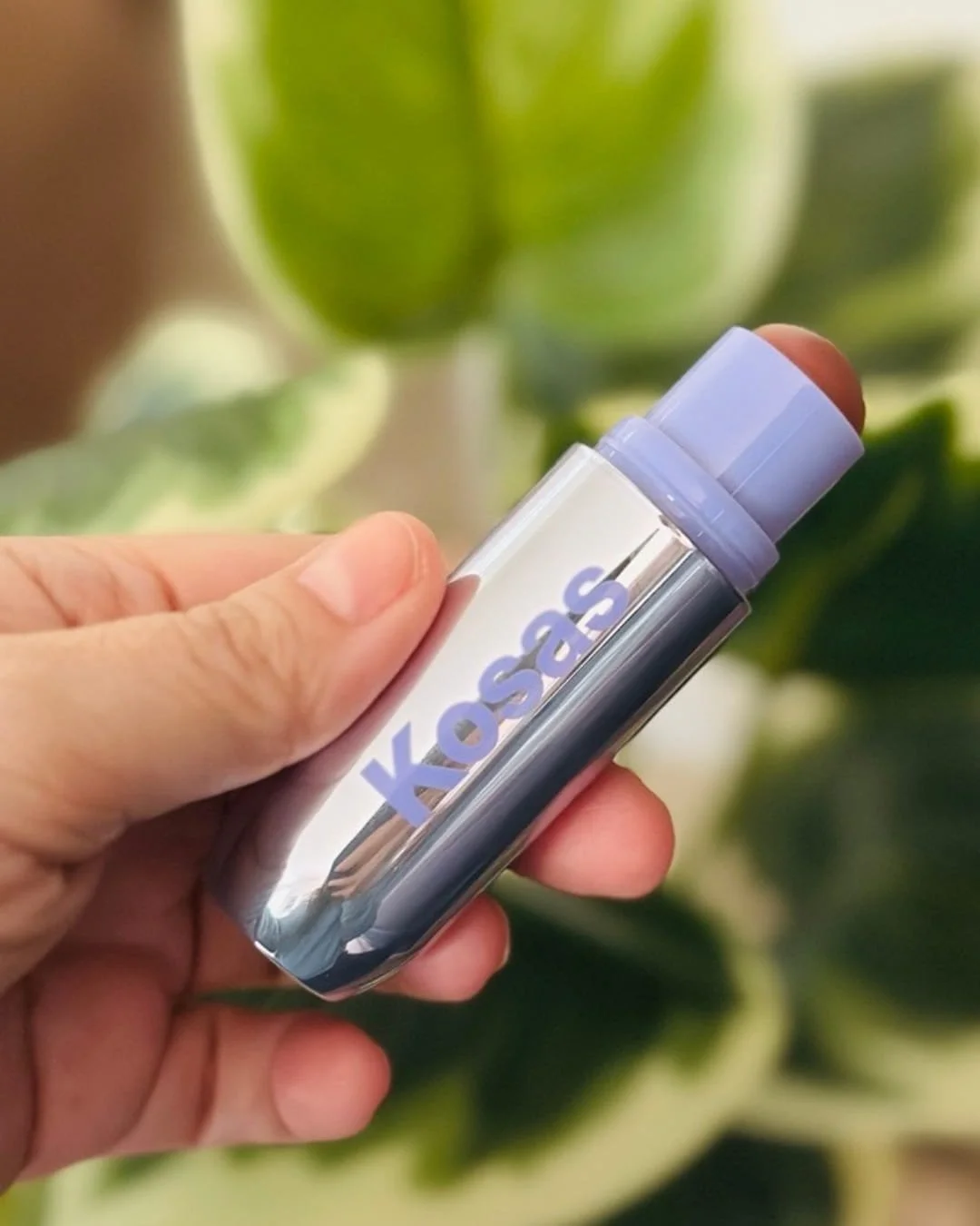 Hand holding a Kosas Multistick Cream in a reflective silver tube with lavender accents against a leafy green background