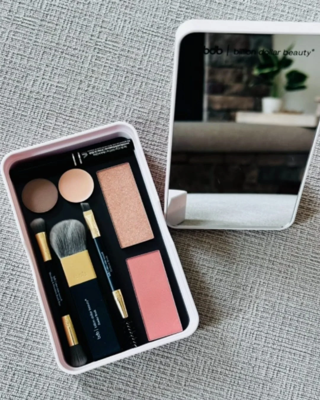 Billion Dollar Beauty Basically Brilliant Bundle featuring a customizable magnetic makeup palette with neutral and warm-toned powders, three black-handled brushes, and a matching white mirror, displayed on a textured gray surface.