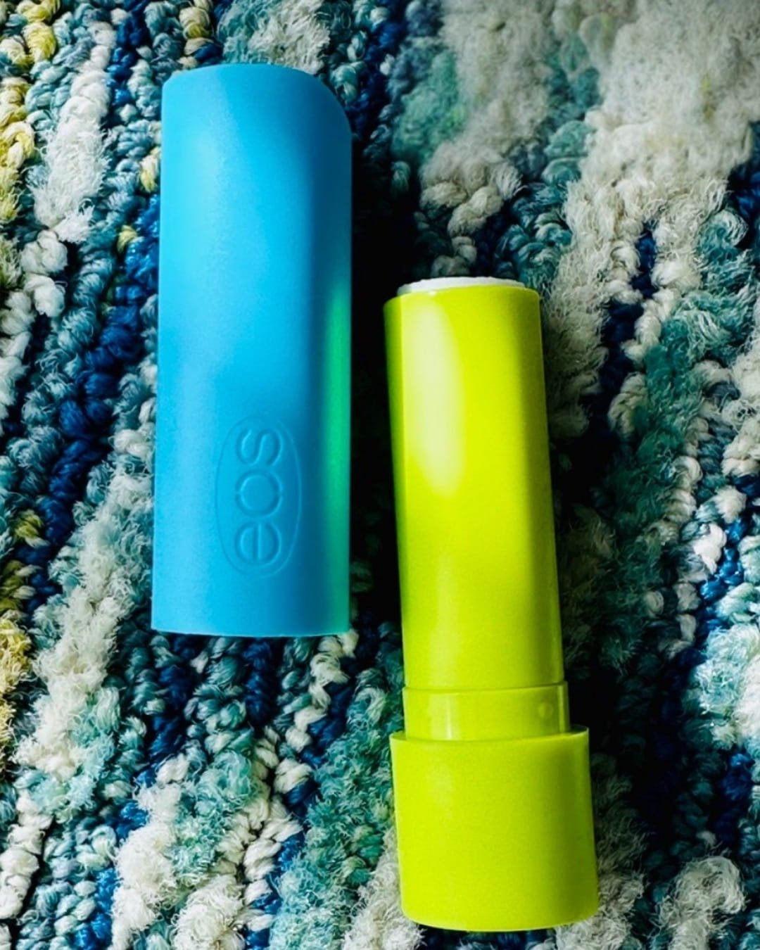 EOS SPF 15 lip balm with a vibrant cyan cap and neon lime green tube, displayed on a textured multicolored rug with woven loops in shades of blue, teal, white, and yellow.