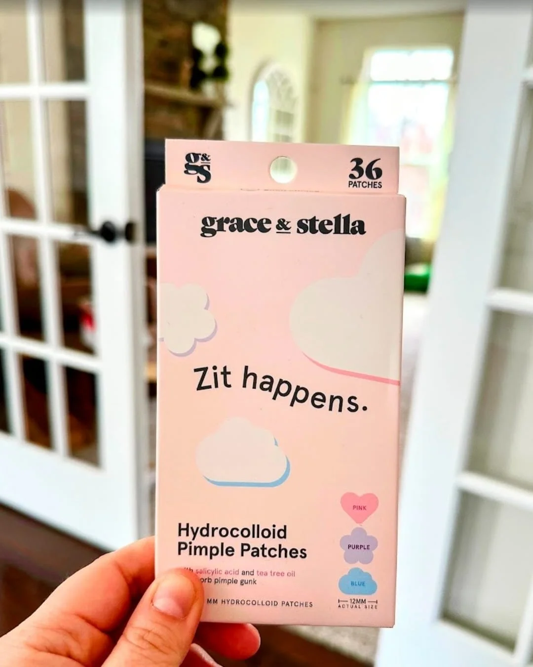Light pink box of Grace & Stella Hydrocolloid Pimple Patches with playful shapes like hearts, flowers, and clouds, featuring 36 patches with salicylic acid and tea tree oil, held in a hand against a blurred indoor background.