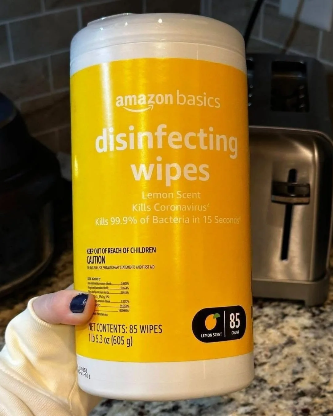 Container of Amazon Basics disinfecting wipes in lemon scent with bright yellow label held in a kitchen setting