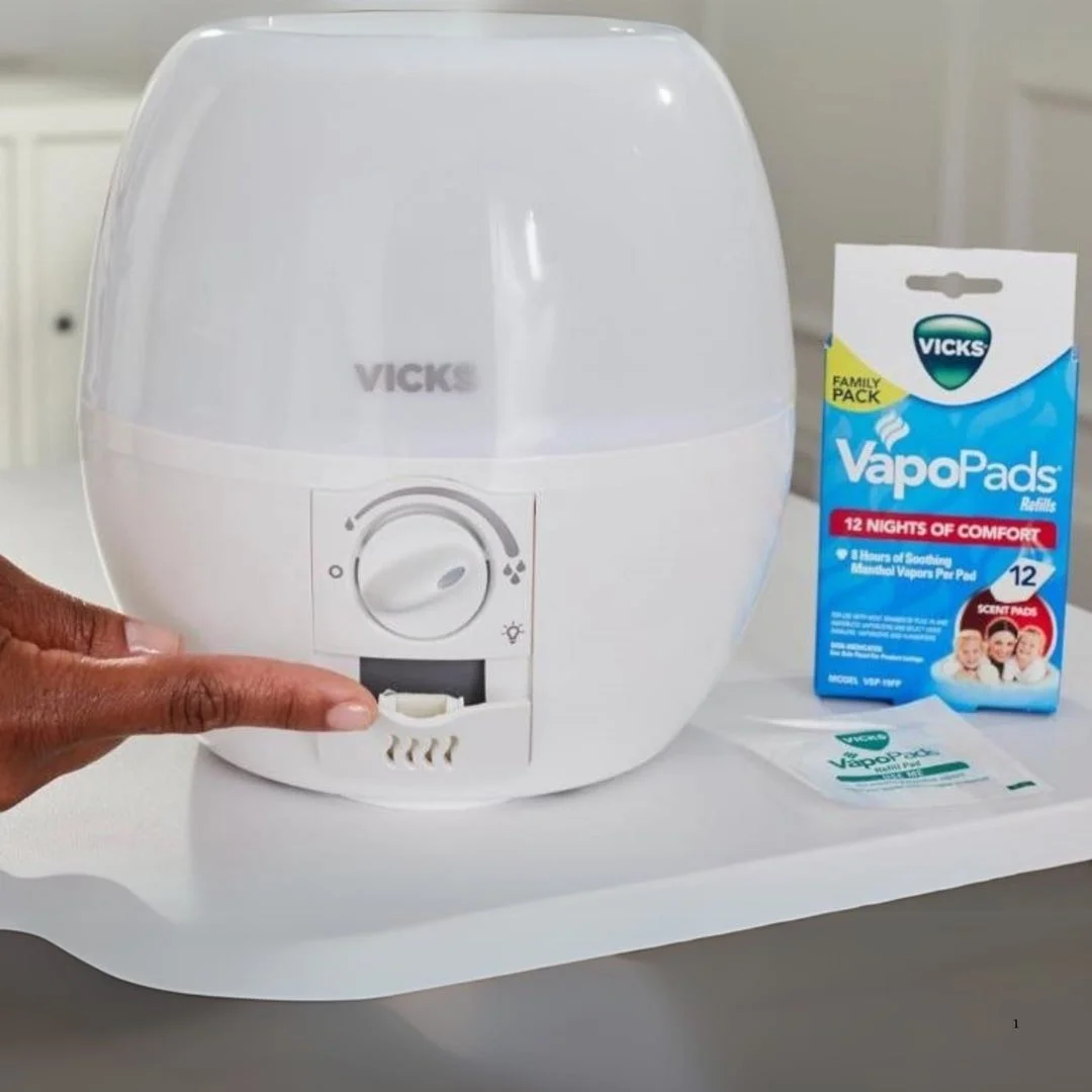 Vicks humidifier with a control dial and a hand adjusting the VapoPads slot, displayed alongside a box of VapoPads Refills and a single refill pad.