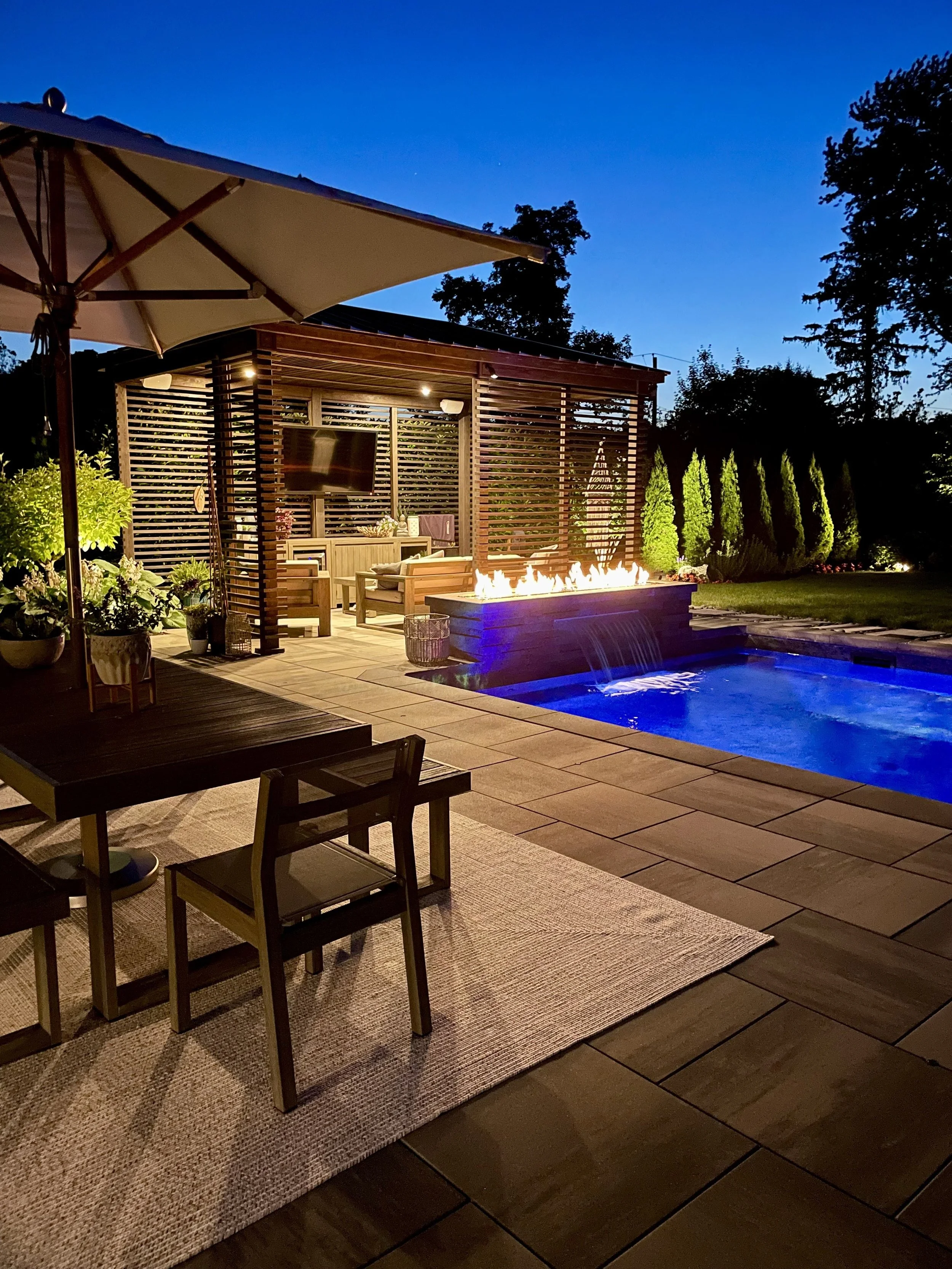 Outdoor backyard patio at night with a swimming pool, fire feature, seating area, large umbrella, and lush trees in the background.