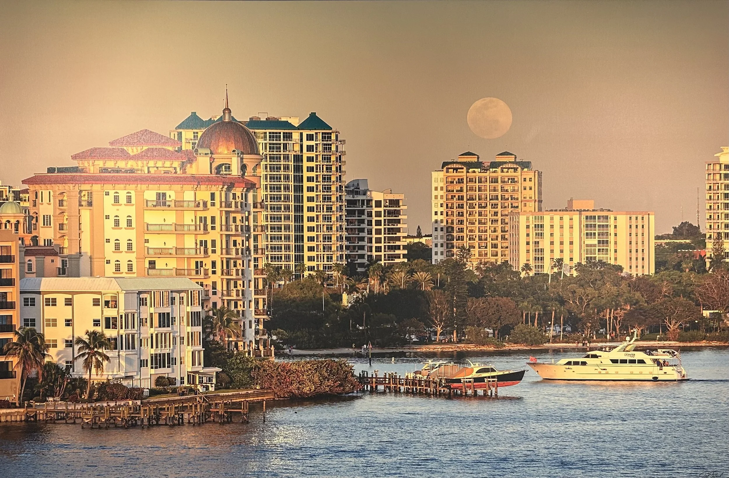 A city skyline at dusk with buildings, a full moon, and boats on a river.