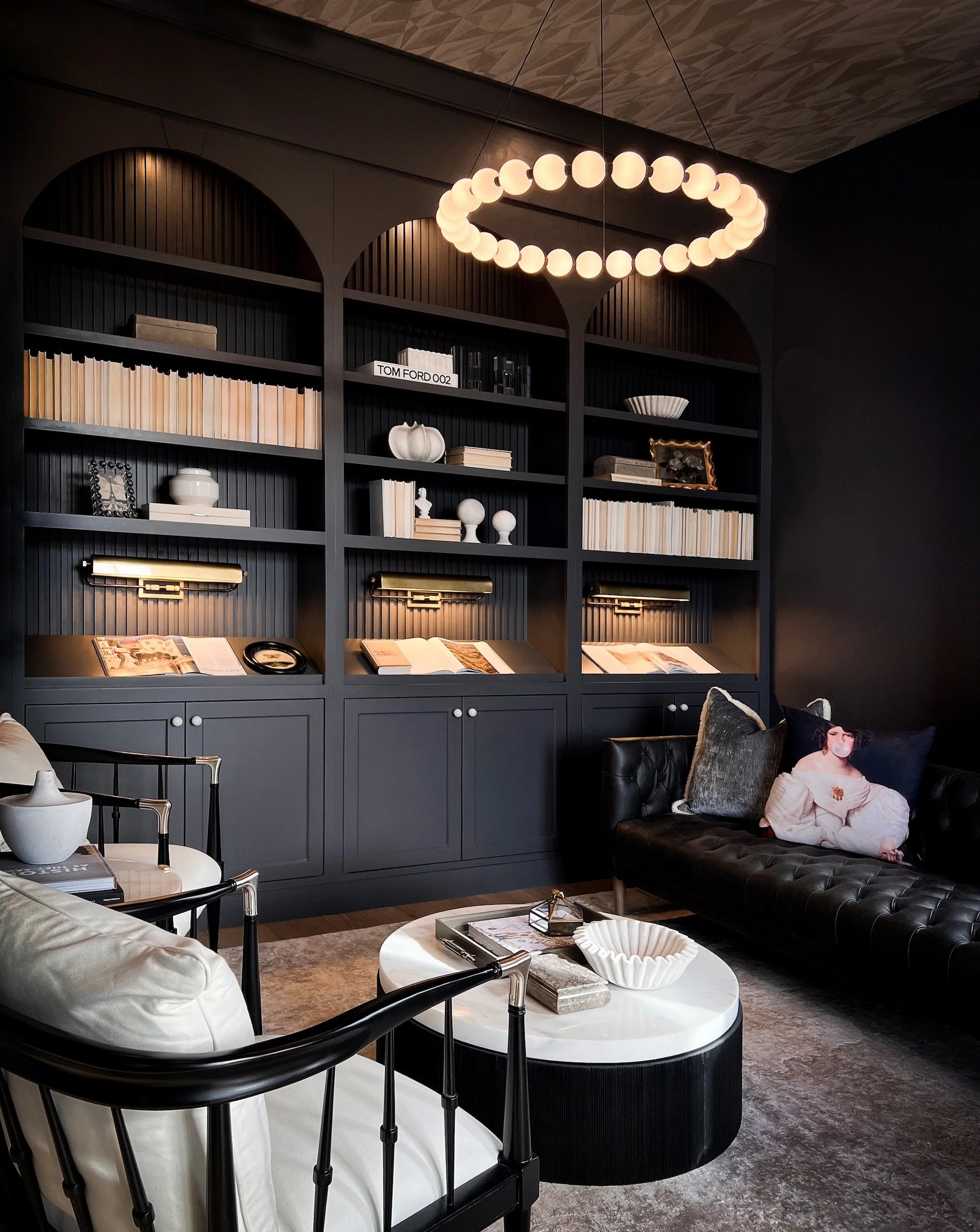 Modern living room with built-in dark blue bookshelf, black leather sofa with decorative pillows, round marble top coffee table, and contemporary chandelier with white globe lights.