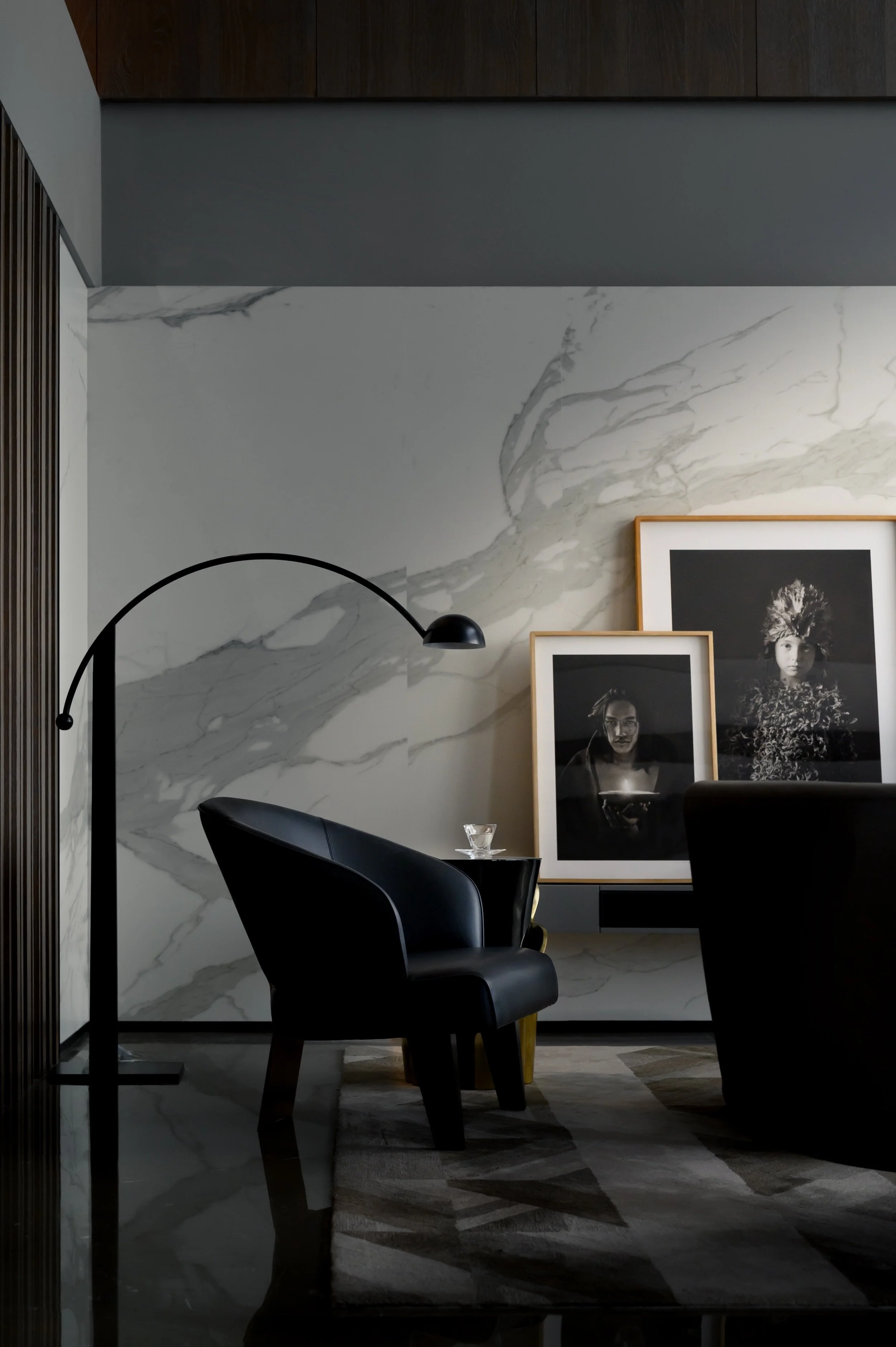 A modern interior with a black leather armchair, an arched floor lamp, and framed black-and-white photographs on a marble wall.