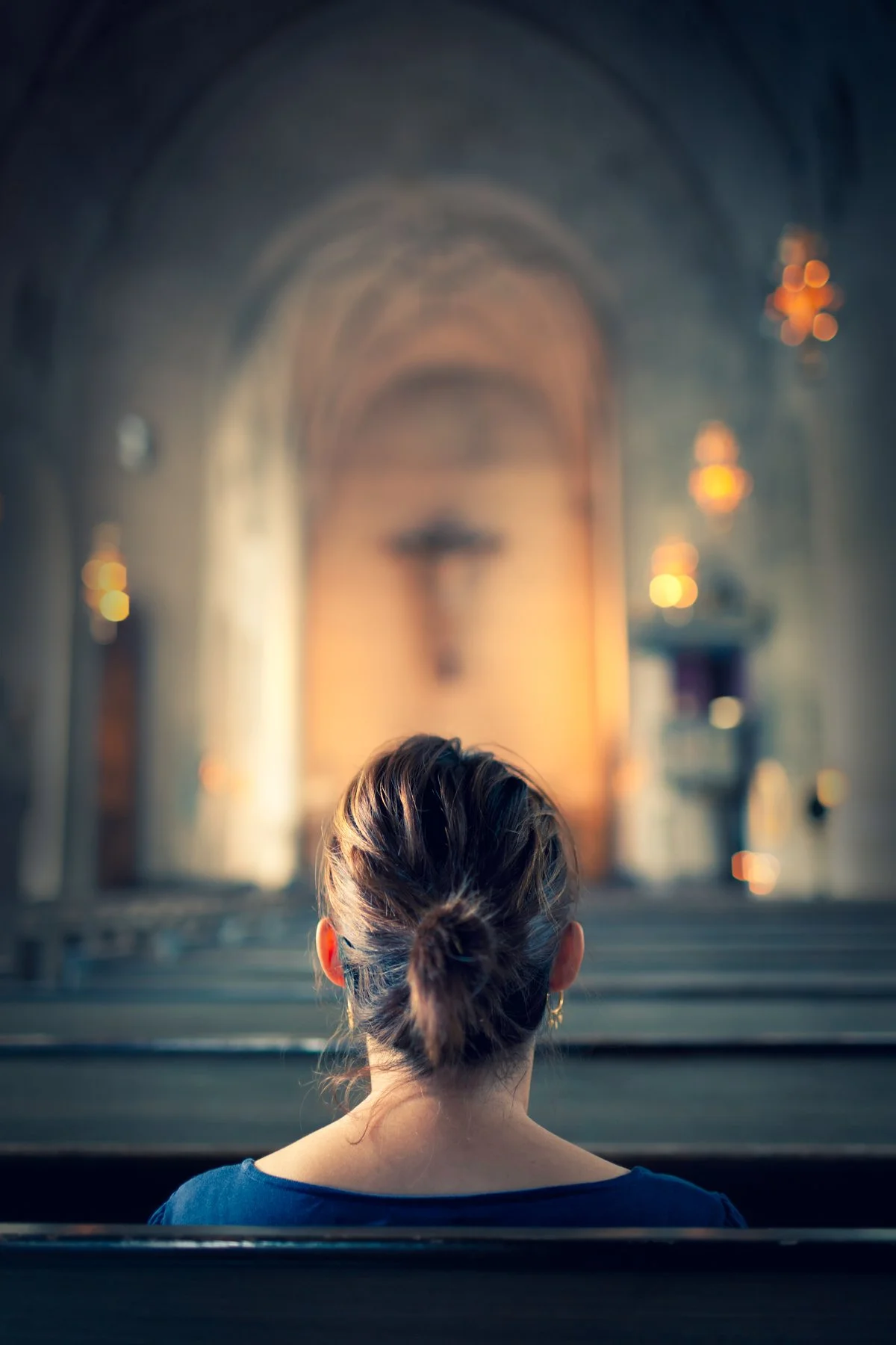 A woman with brown hair tied in a bun, sitting alone in a church pew, facing the altar with a crucifix, candles, and religious decorations in the background.