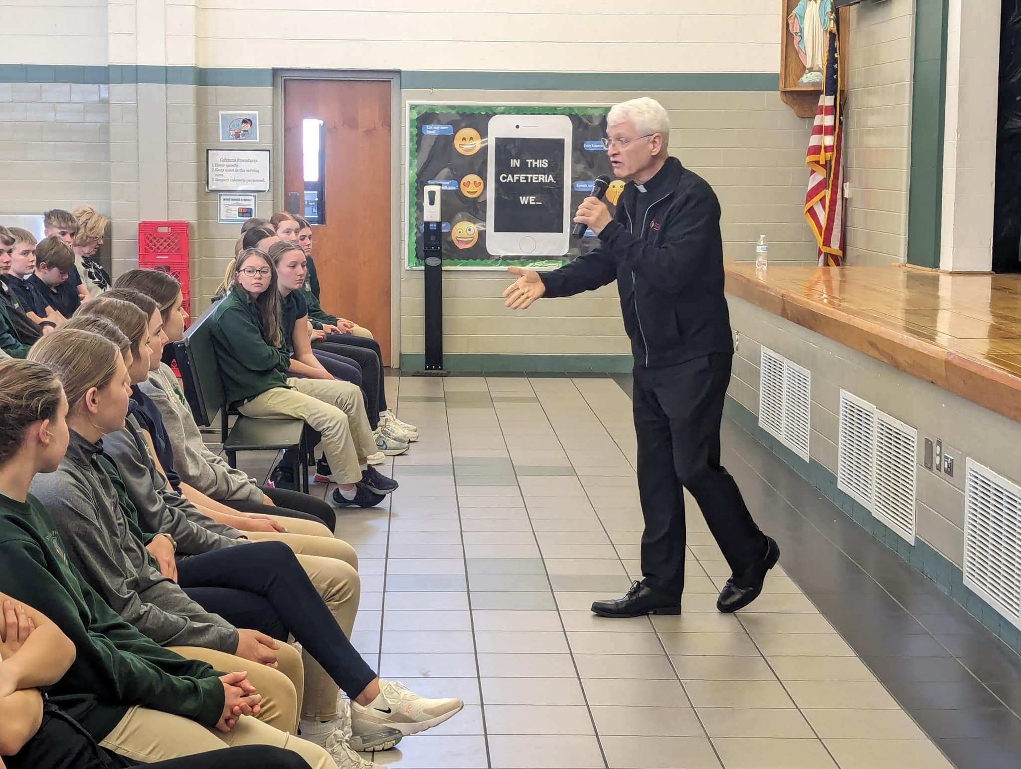A man speaking to a group of students seated in a school cafeteria.