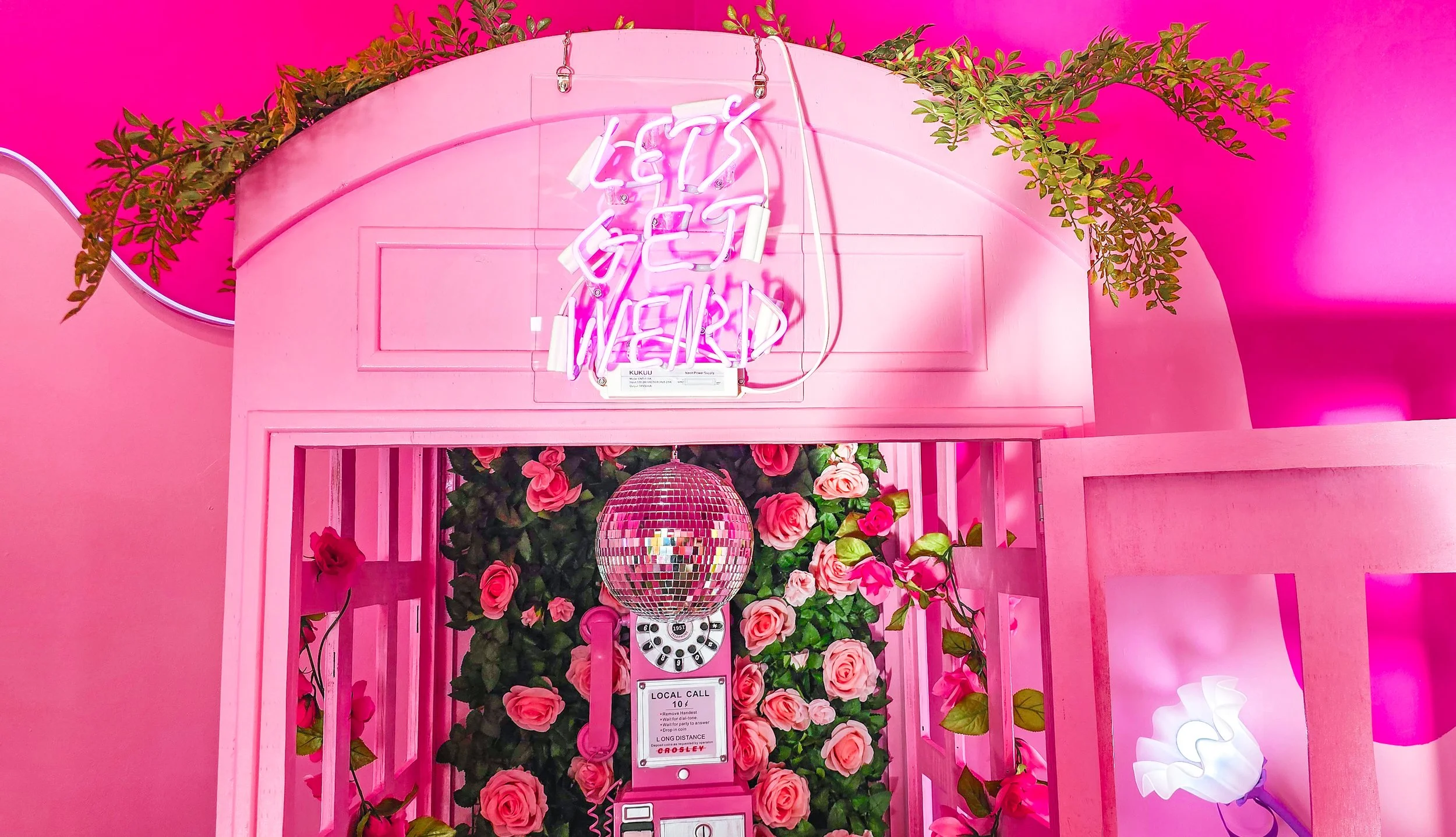 Pink telephone booth decorated with pink roses, a disco ball, a neon sign that reads 'Let's Get Married', and neon lighting creating a vibrant, romantic atmosphere.