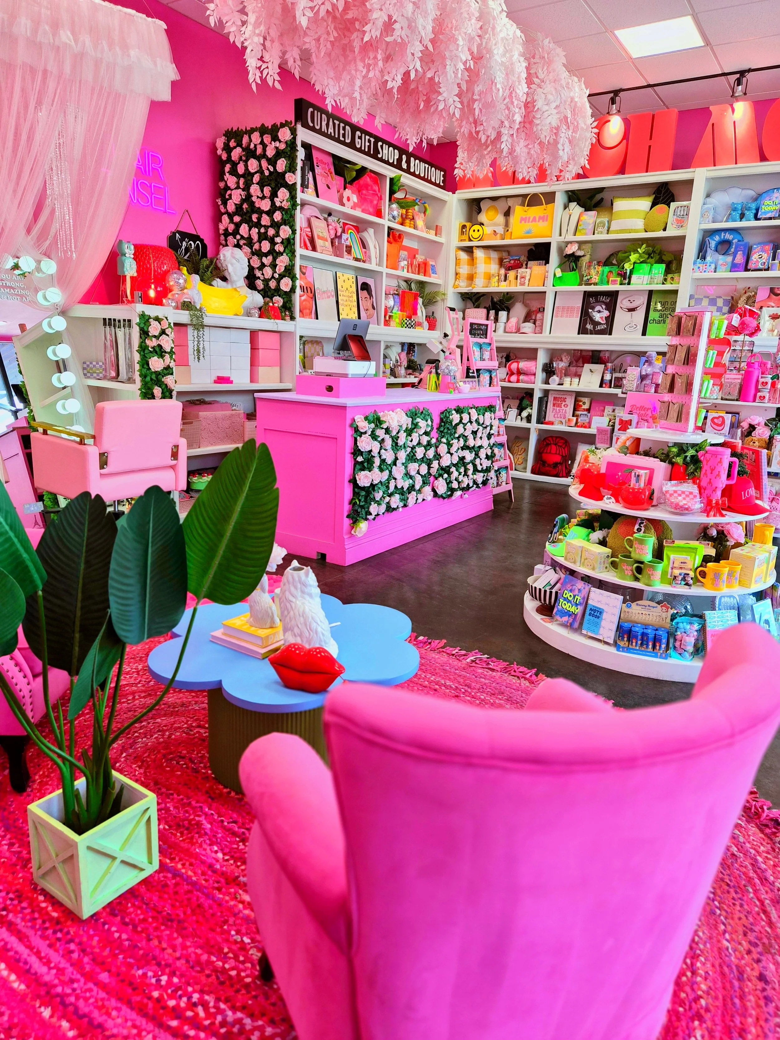 A colorful gift shop decorated with pink and white accents. Pink armchair and a blue cloud-shaped table with a red lips sculpture and white animal decor. Shelves filled with various gift items, books, and toys. Pink floral arrangements, greenery, and hanging white leaves decor. Bright lighting and a pink textured rug.