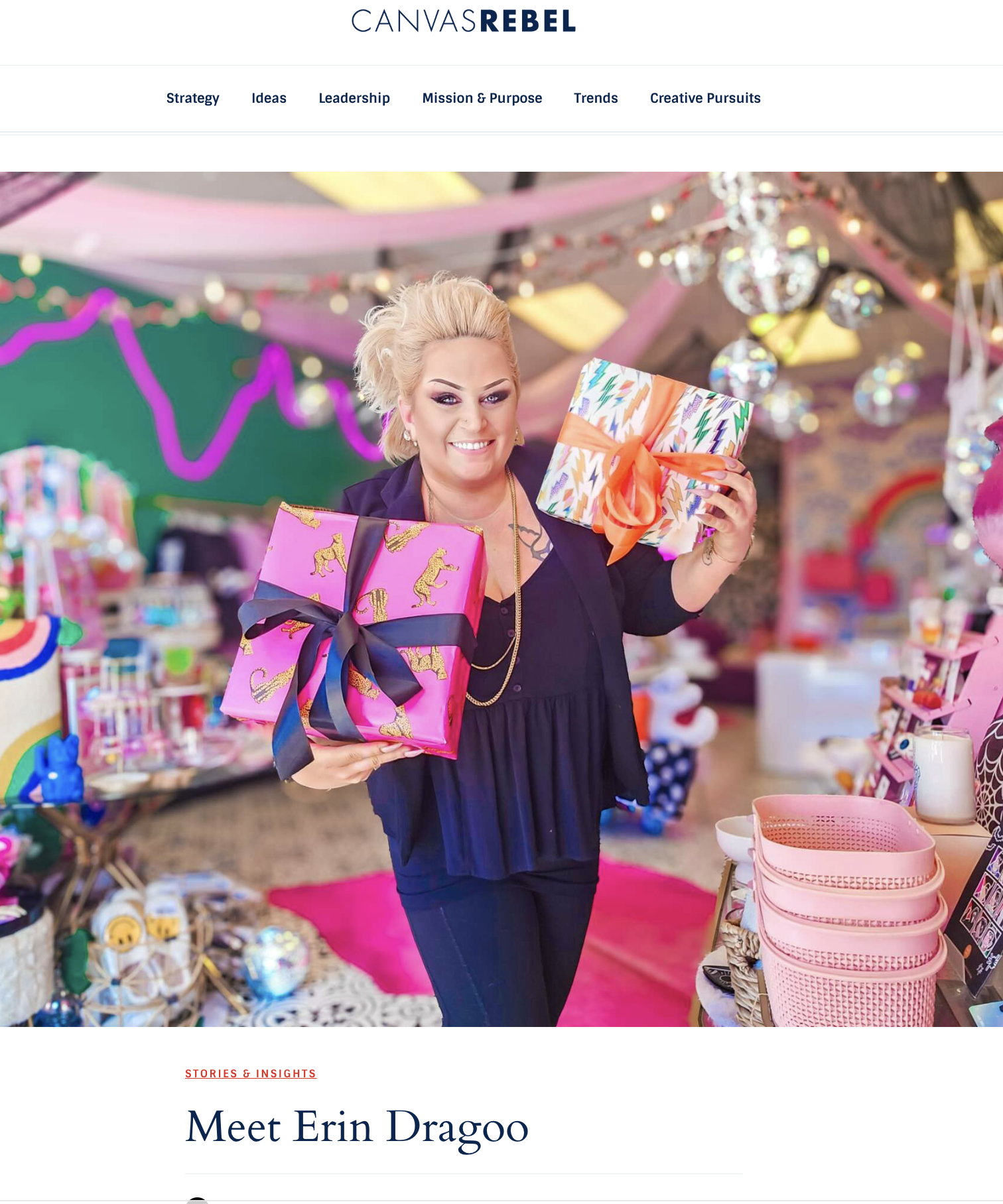 A woman with short blonde hair, smiling, holding colorful wrapped gifts with bows inside a festive, decorated store with pink and multicolored decor.