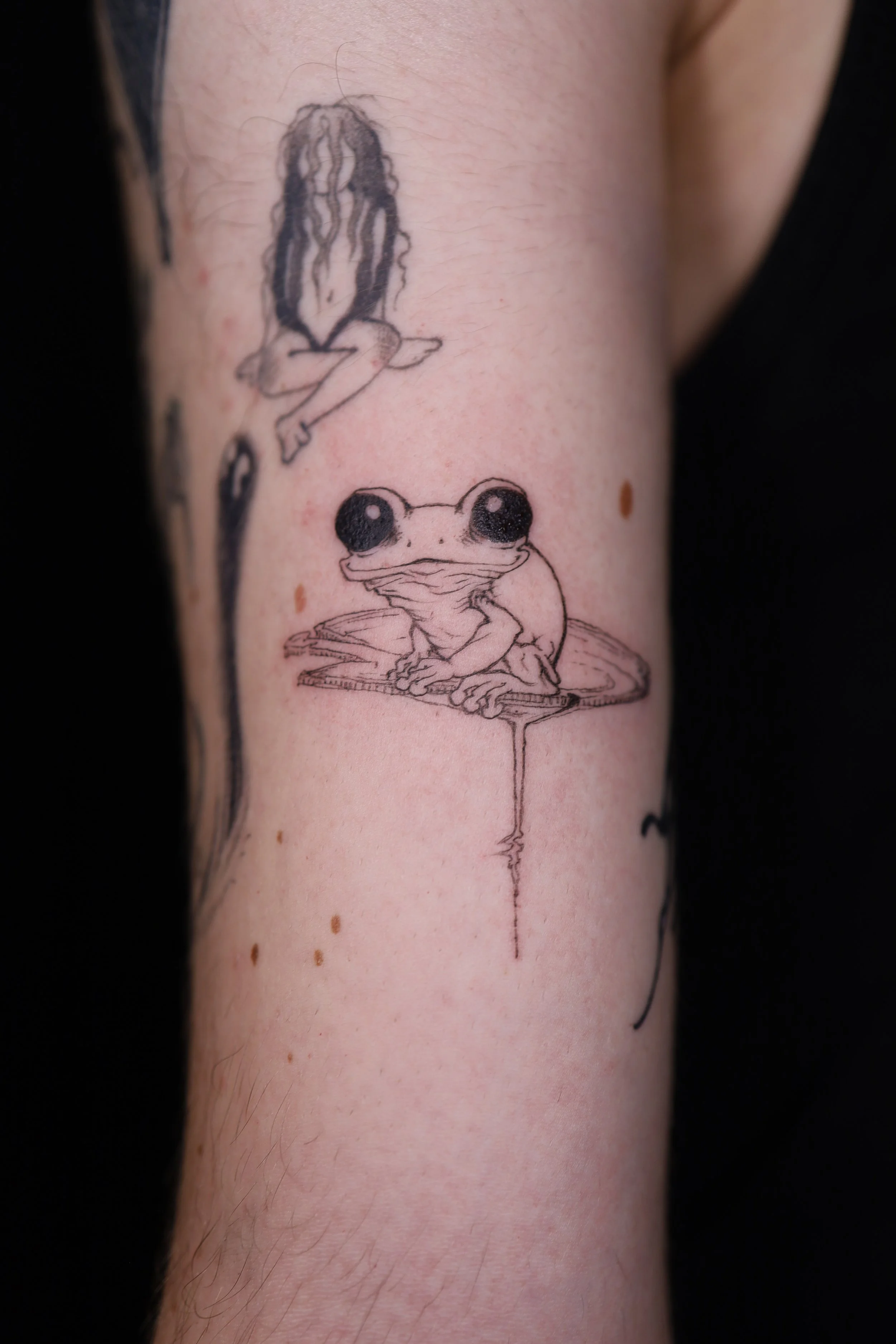 Tattoo of a frog with large eyes sitting on a leaf, with a girl sitting cross-legged above it, on a person's arm.