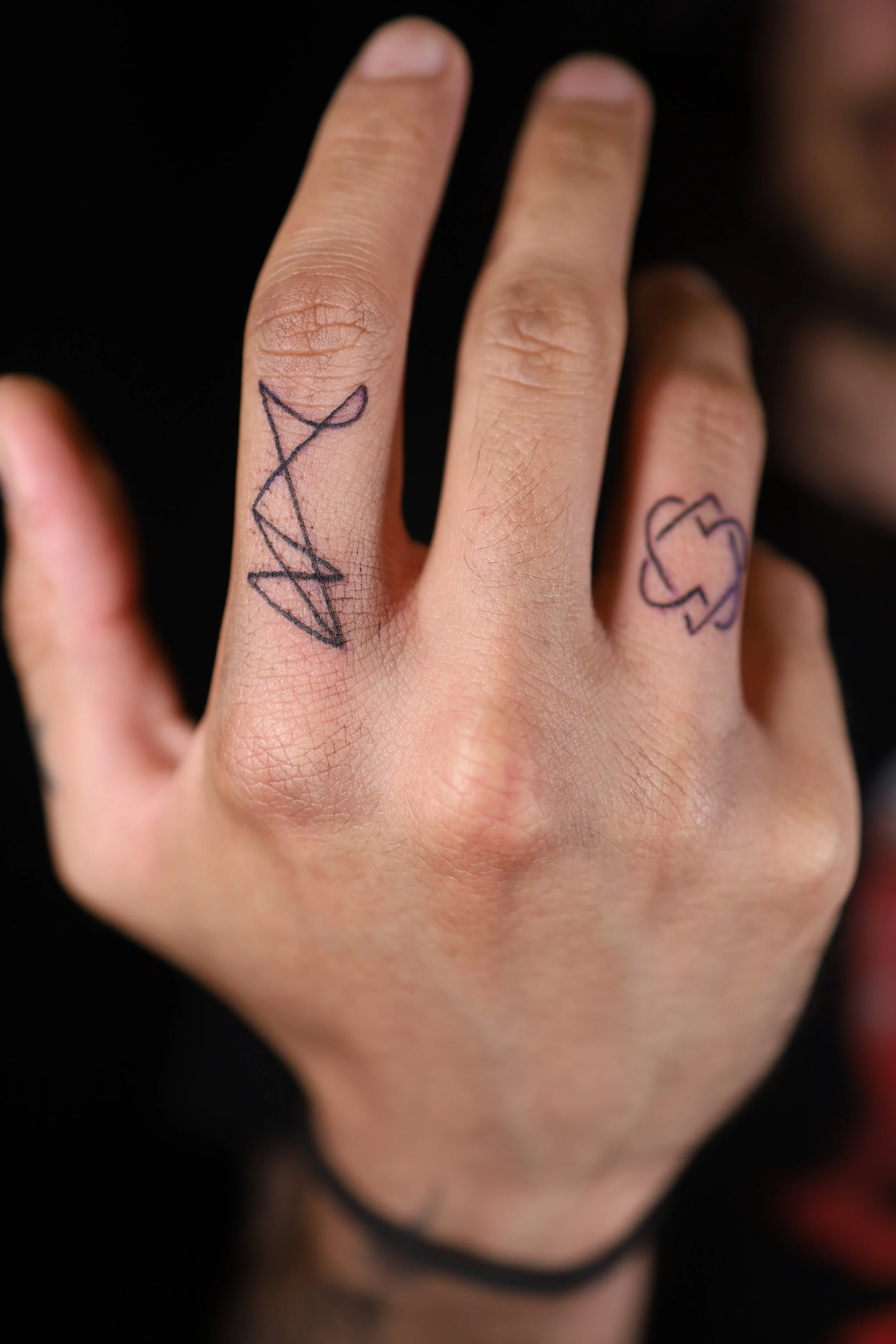 Close-up of a person's hand showing two tattoos on the middle and ring fingers. The tattoo on the middle finger is a black geometric triangle design, and the tattoo on the ring finger is a black outline of a dollar sign.
