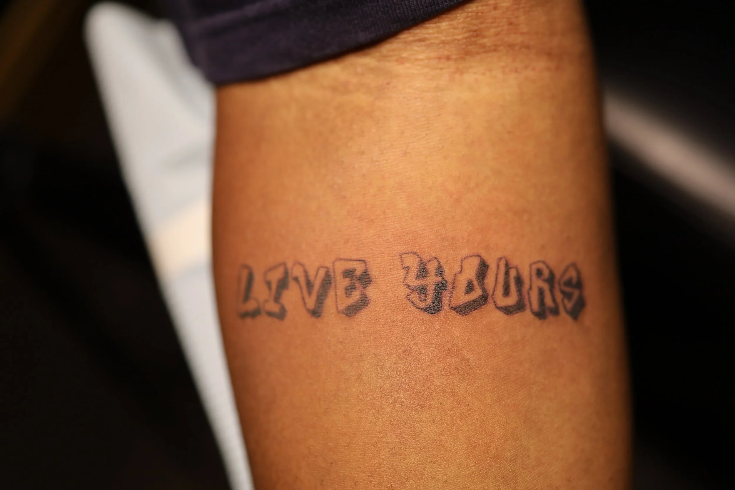 Close-up of a person's arm with a tattoo that reads 'LIVE 4 US' in bold, stylized letters.