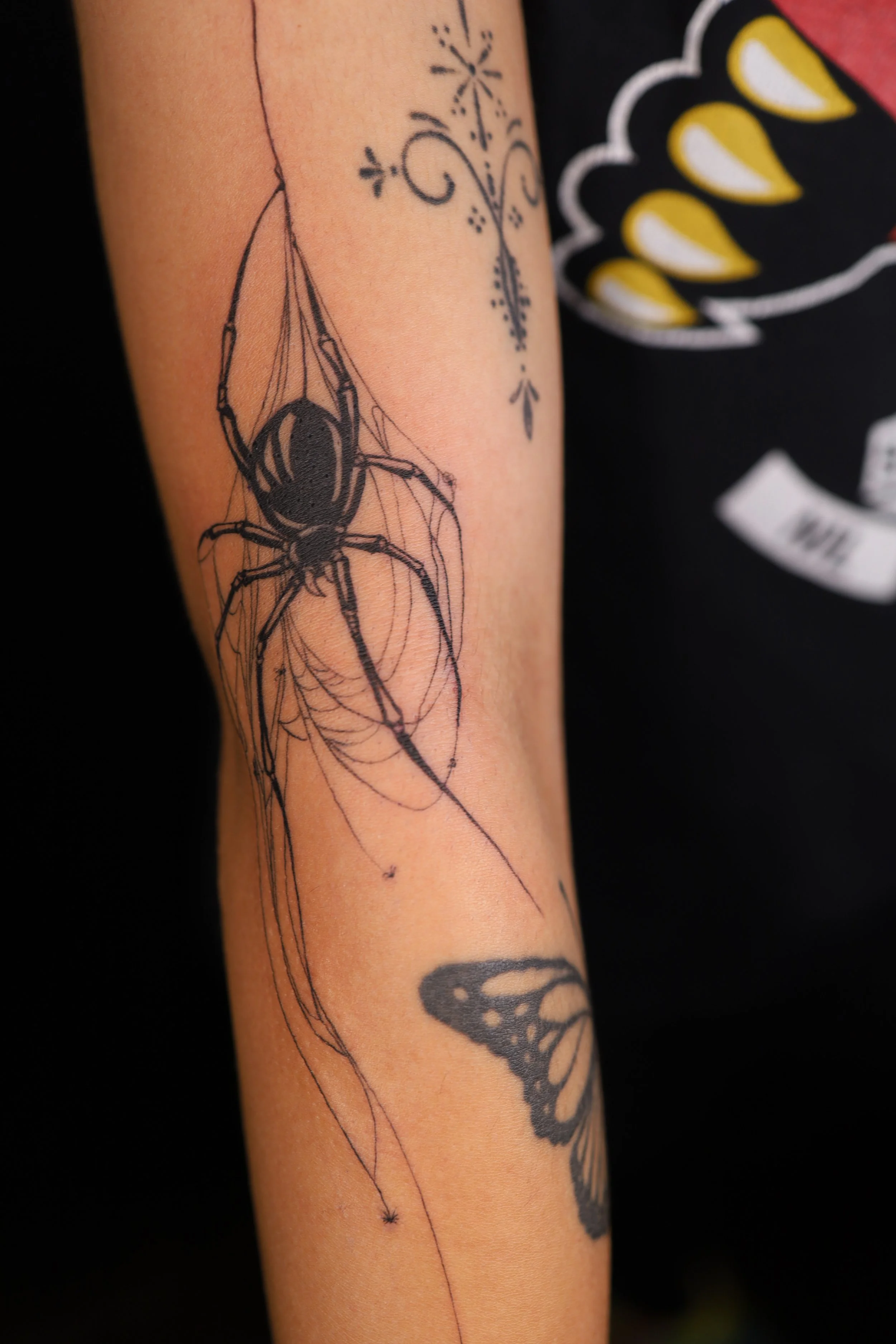 Tattoo of a spider on the arm, with a butterfly tattoo below and decorative line work above.