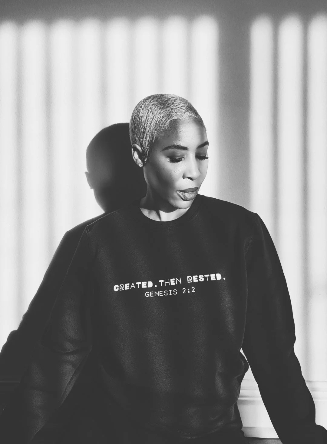 A black woman with short blonde hair wearing a black sweatshirt with the words 'Created. Then Rested. Genesis 2:2' printed on it, standing against a wall with shadows cast from vertical blinds.