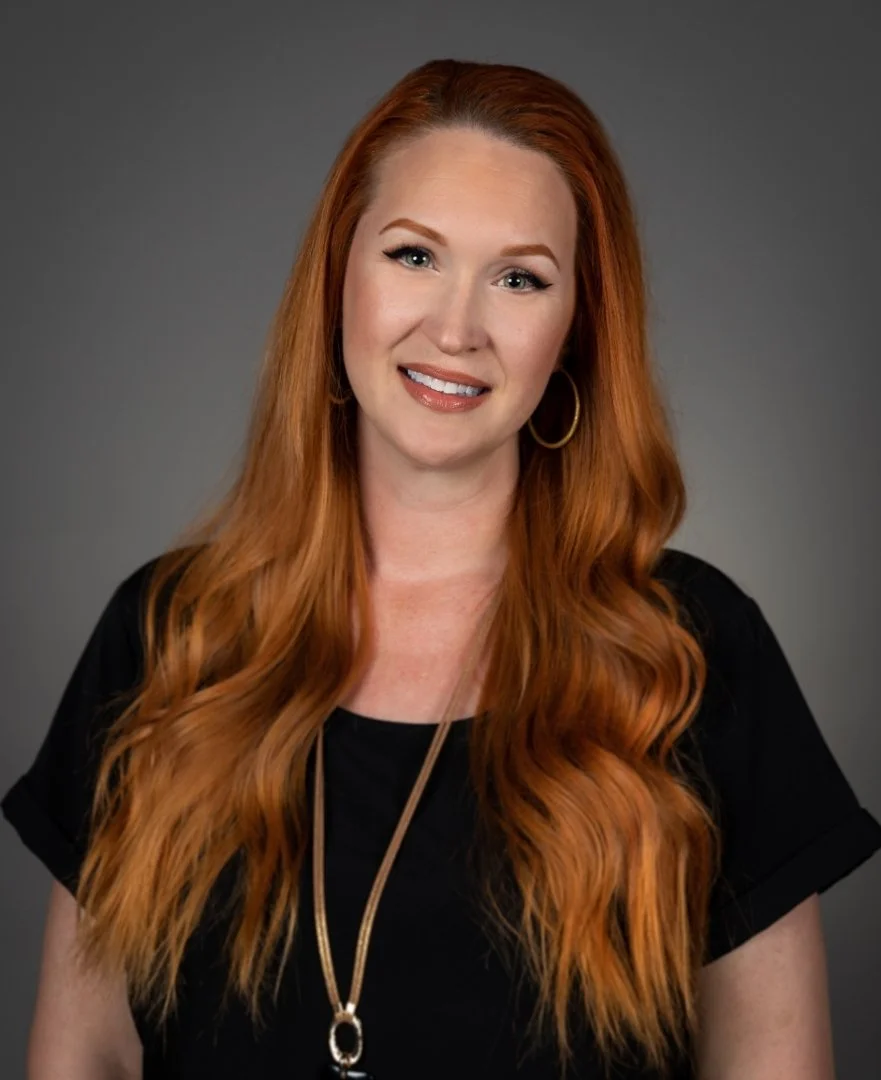 Smiling woman with long red hair, wearing a black top and gold hoop earrings, against a gray background.