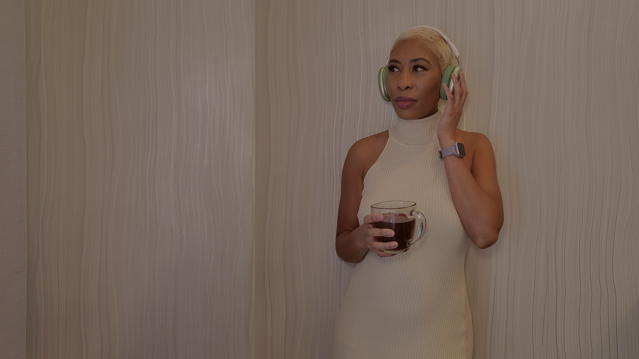 Woman with blonde hair in a sleeveless beige turtleneck dress holding a glass of dark beverage and wearing a smartwatch on her left wrist, standing against a textured light-colored wall, touching her large two-toned headphones with her right hand.