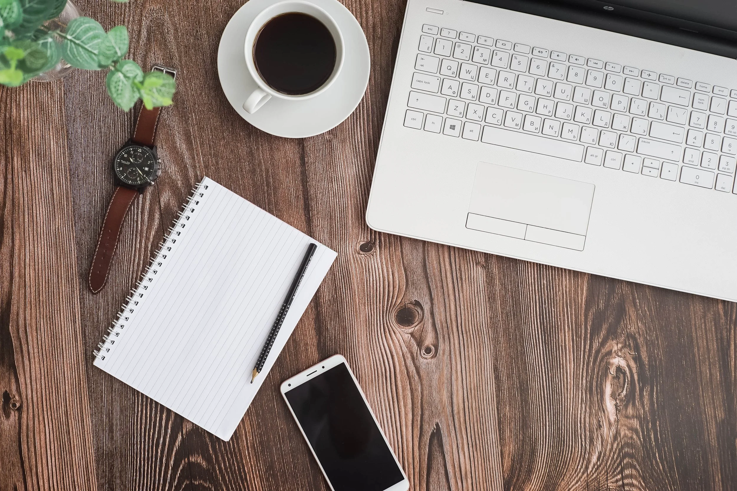 Top-down view of a wooden desk with a laptop, coffee cup, notebook with pen, smartphone, wristwatch, and a potted plant.