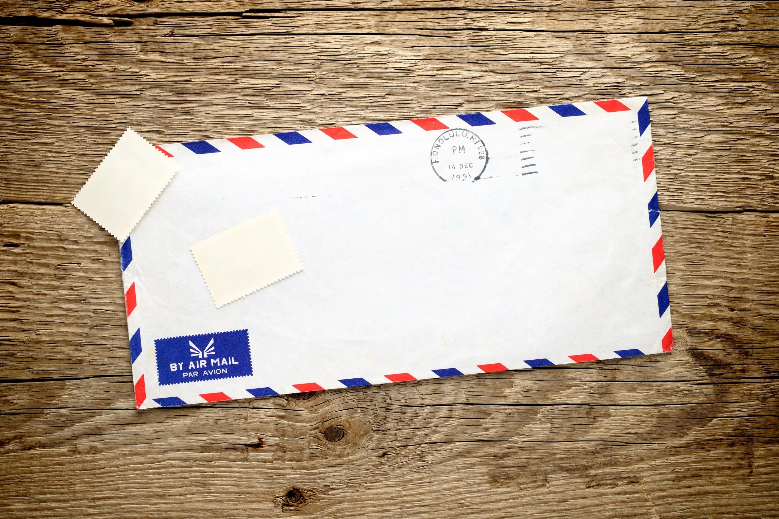 A blank airmail envelope with red and blue border, two blank stamps, and a sticker that says 'BY AIR MAIL' on a wooden surface.