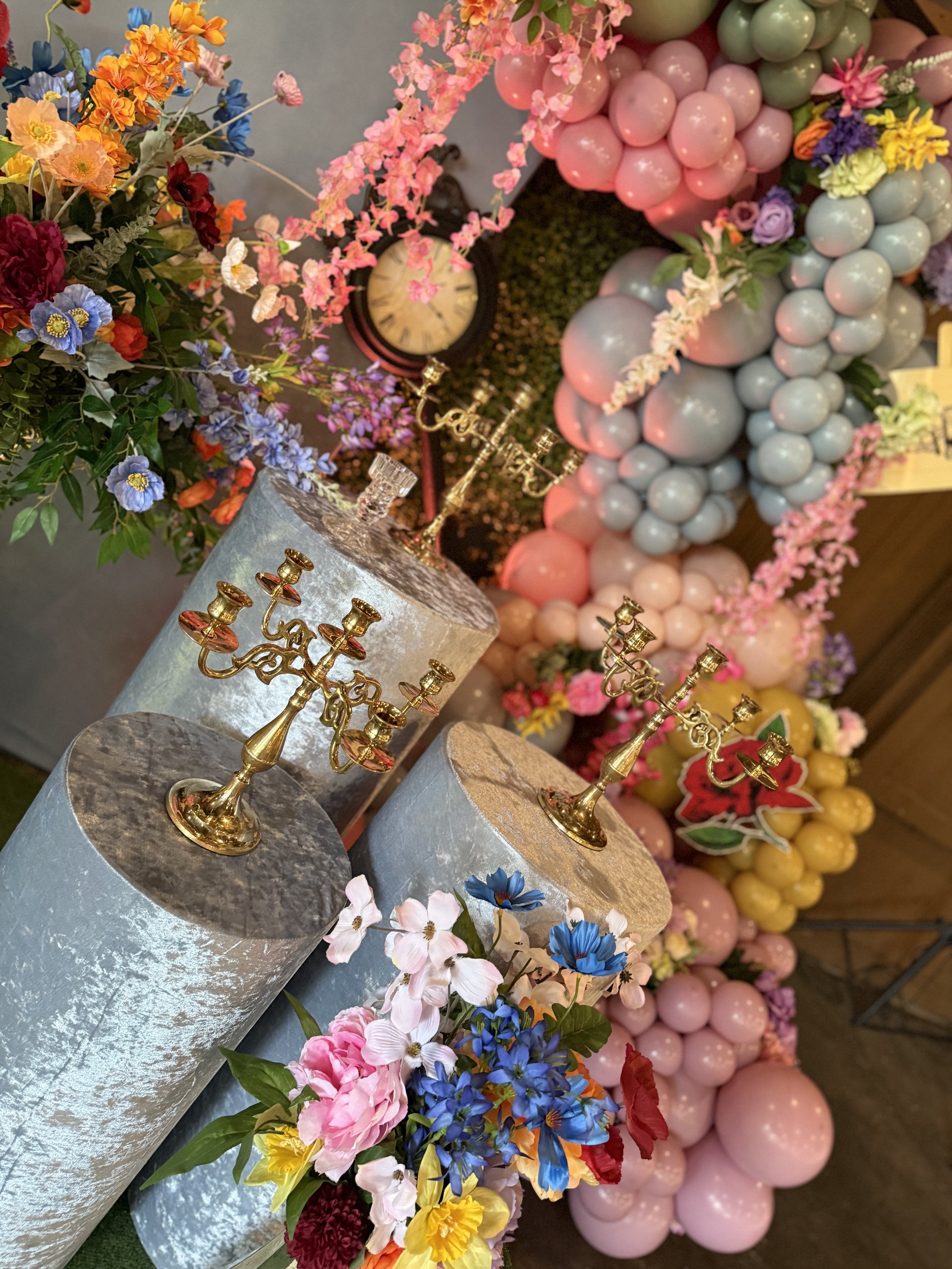 Decorative floral arrangement with pink, blue, red, and yellow flowers, balloons in pastel pink, green, and yellow, and gold candelabras on metallic pedestals. A clock with pink flowers in the background.