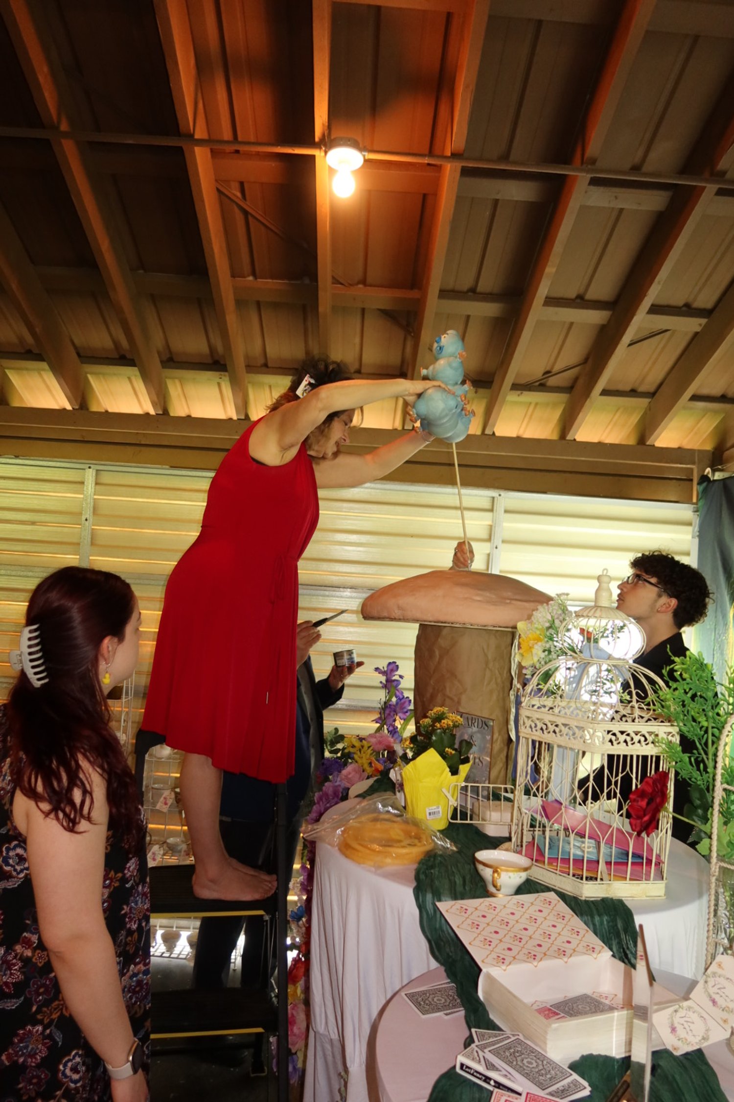 Woman in a red dress standing on a step stool, holding a puppet, performing a puppet show with a man sitting behind a table decorated with flowers, birdcages, and cards. Two other spectators watch the scene.
