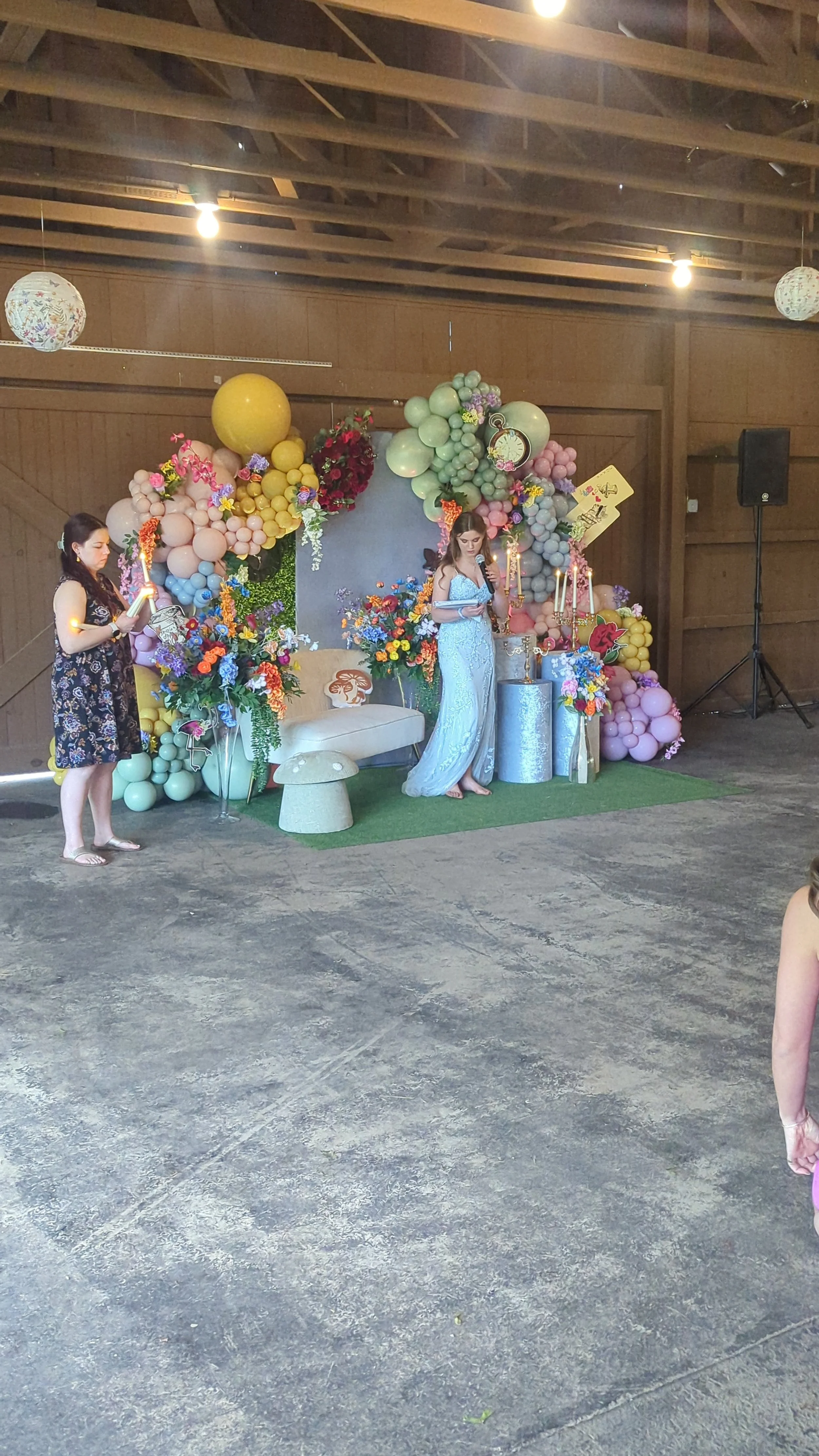 Indoor celebration with floral and balloon decorations, two women with one reading from paper and the other holding a microphone, on a decorated stage with candelabras and a backdrop.