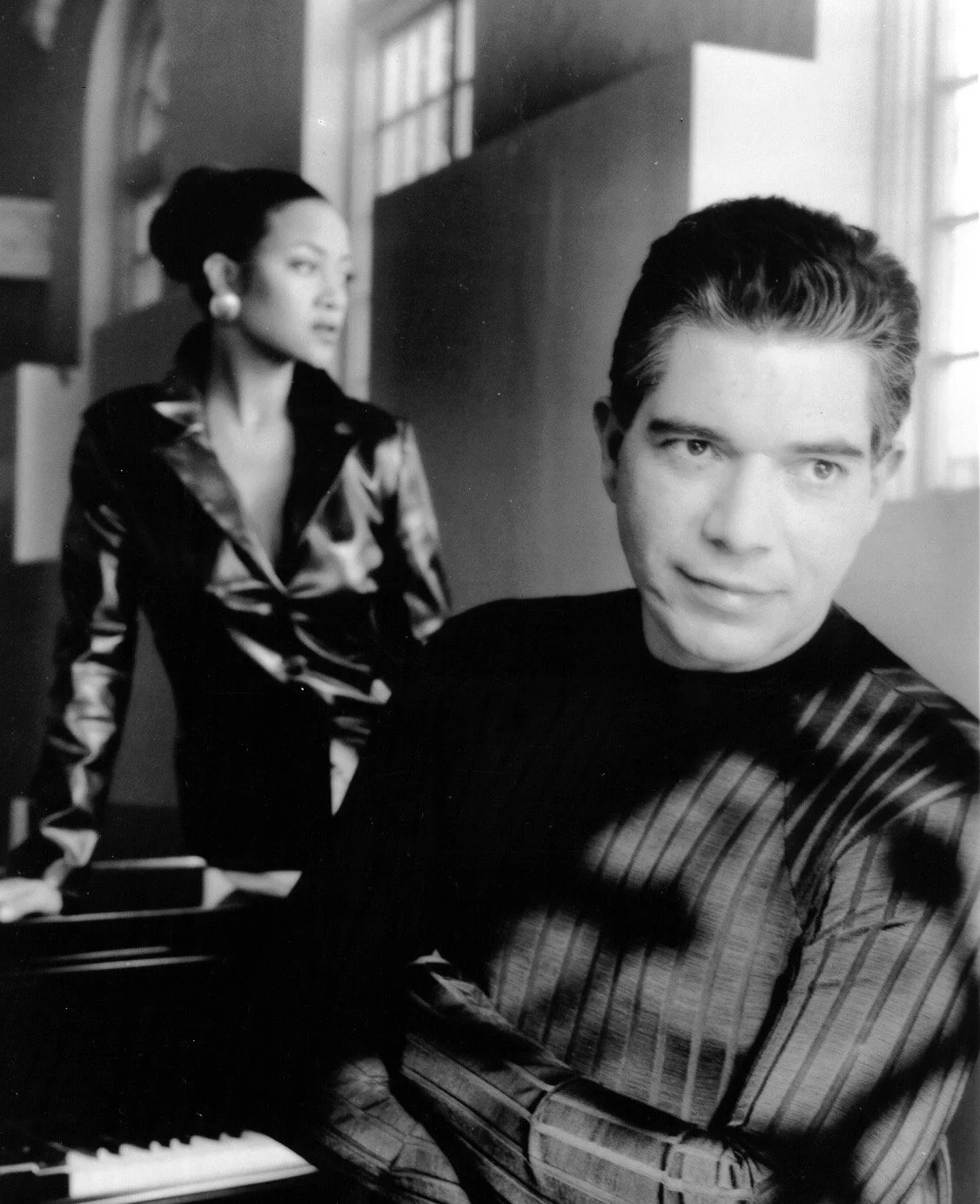 A black and white photo of a man sitting at a piano with a woman standing behind him in a room with large windows.