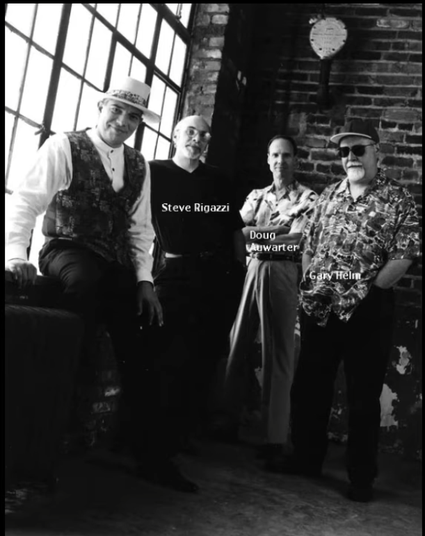 Four men standing and sitting against a brick wall and large window in a loft-style space. They are labeled as Steve Rigazzi, Doug Auwarter, and Gary Helm, with the fourth man not labeled. The photo is in black and white.