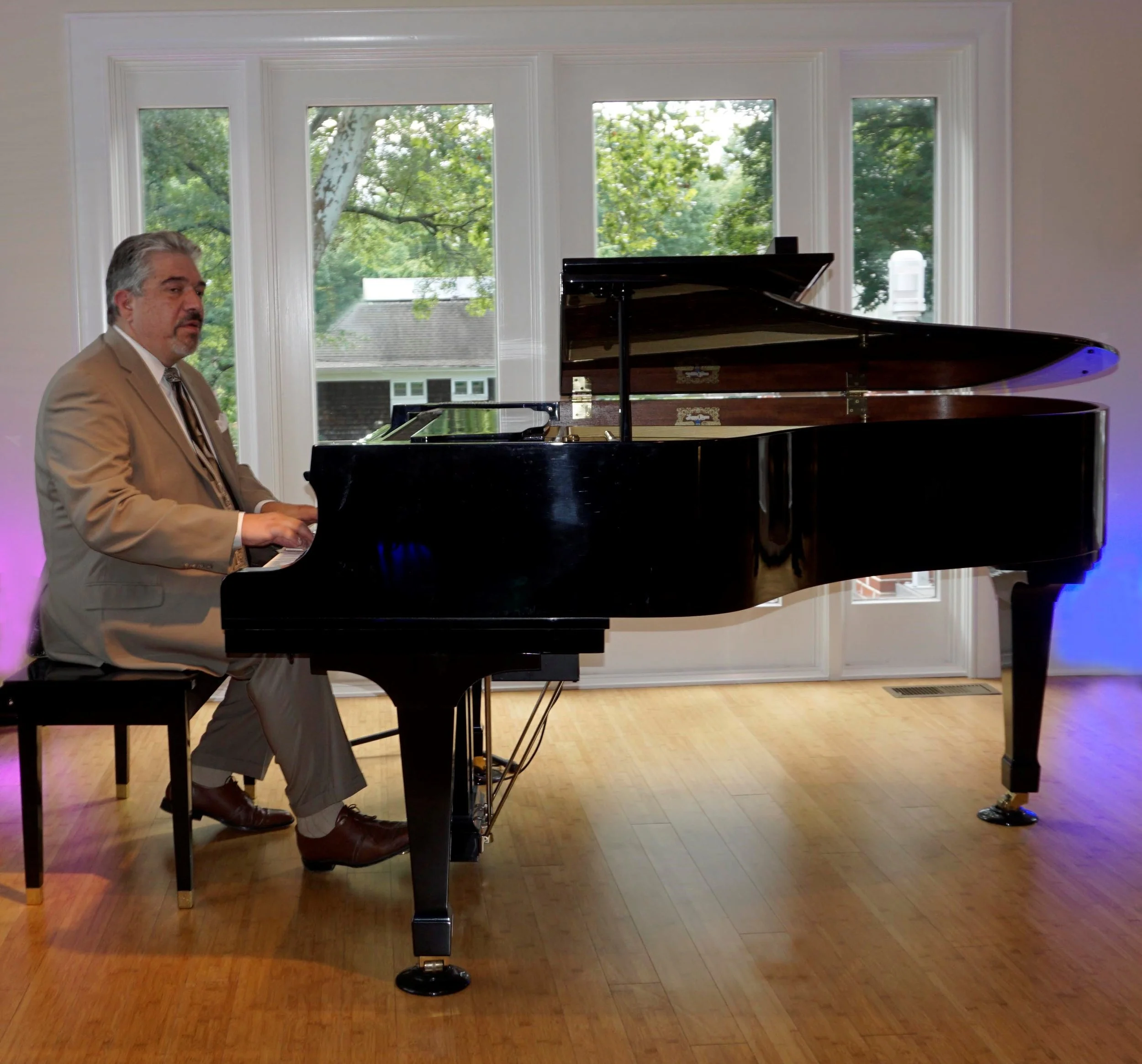 Man in a beige suit playing a black grand piano in a room with large windows showing trees outside.