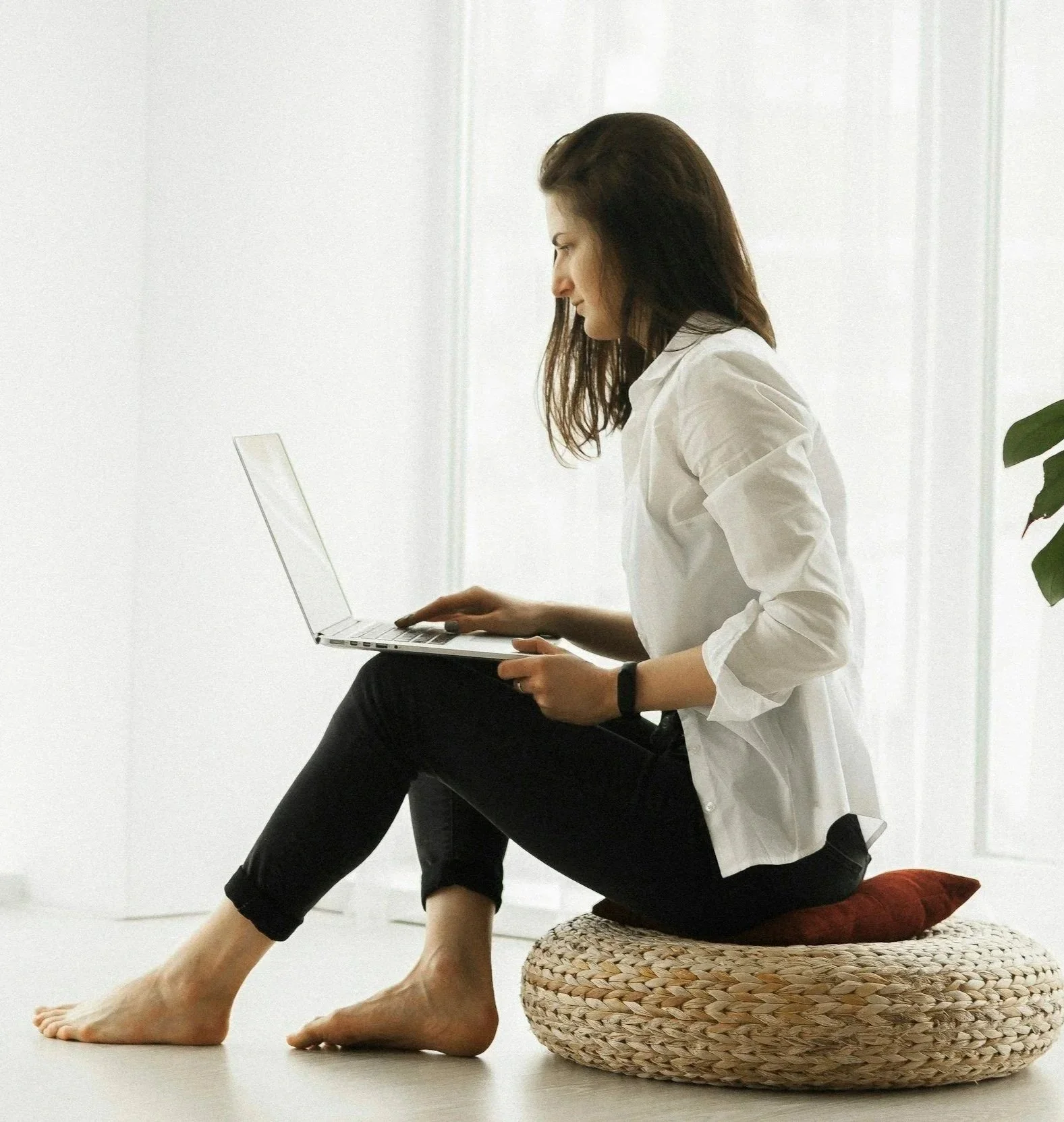 A young woman sitting on a woven pouf with a red cushion, working on a laptop in a bright, minimalistic room.